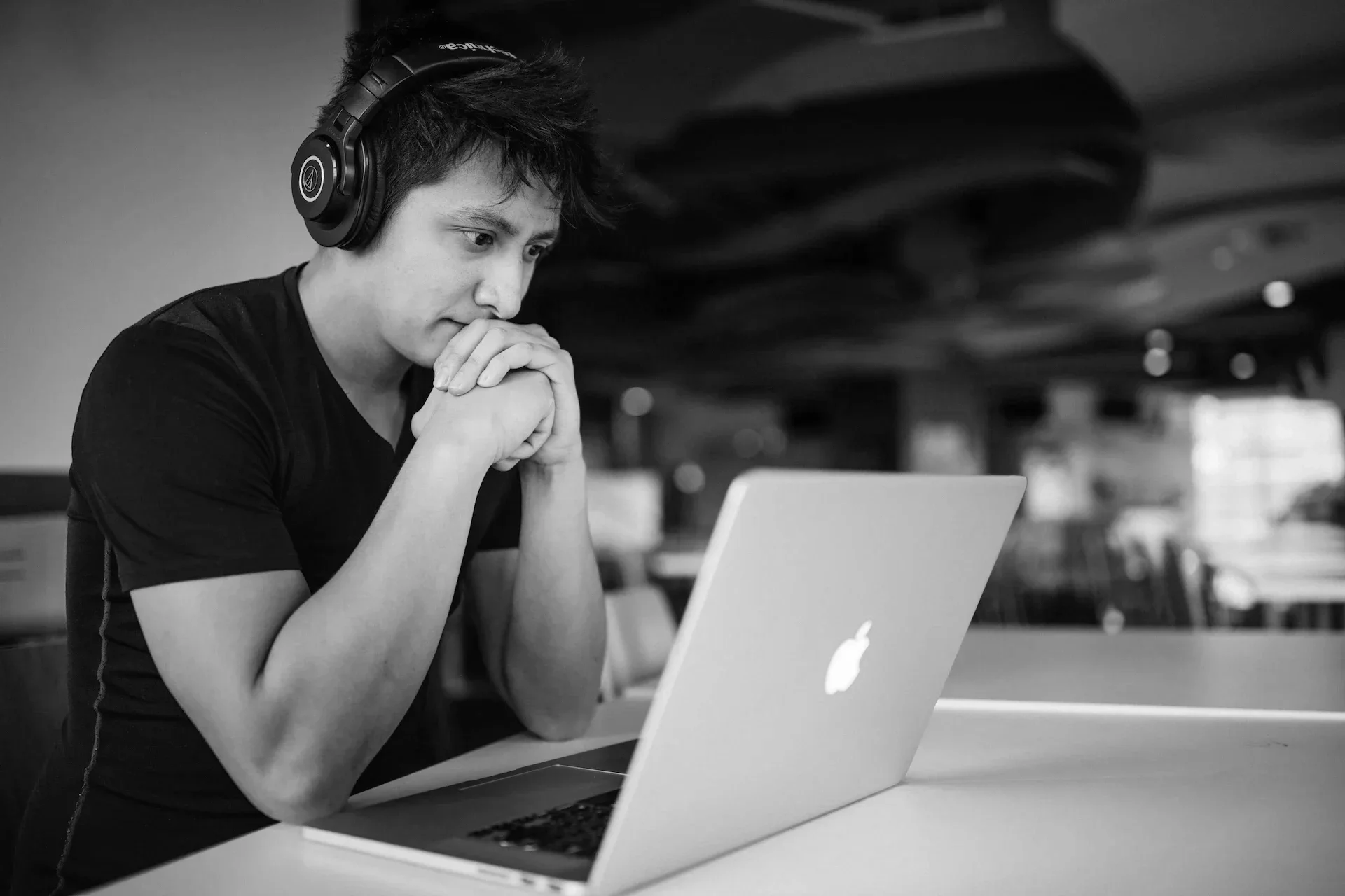 A young man sitting at a table working on a laptop, wearing headphones and appearing focused, in a modern indoor setting.