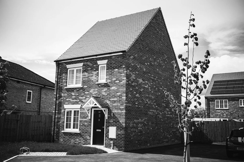 Black and white photo of a modern brick house with gabled roof, white window trims, front door with small porch, small tree in front yard, neighboring houses, fence, and car parked on driveway.