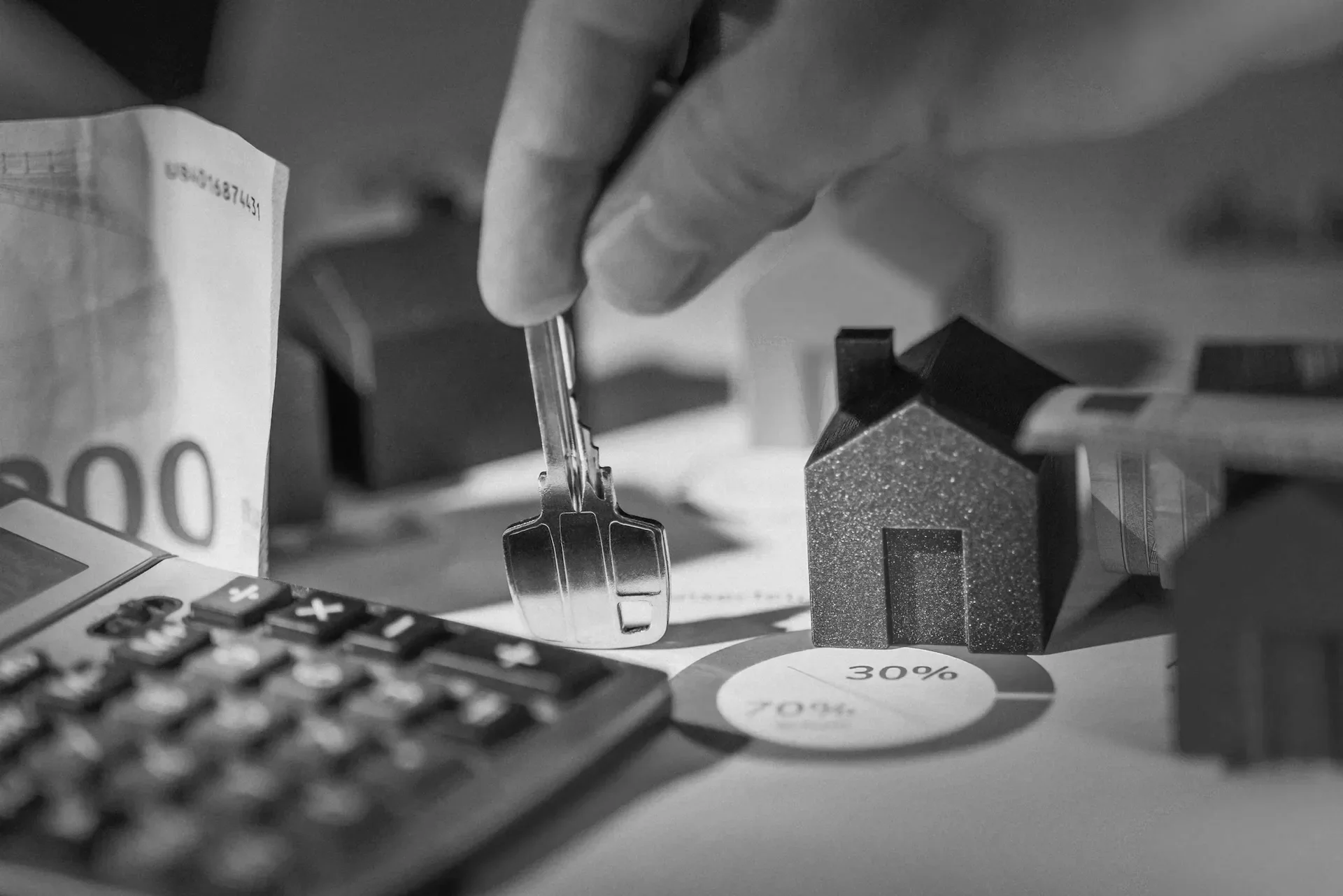 Close-up of a person's hand holding a key over a desk with a calculator, a small house model, and a superimposed pie chart showing 30%.