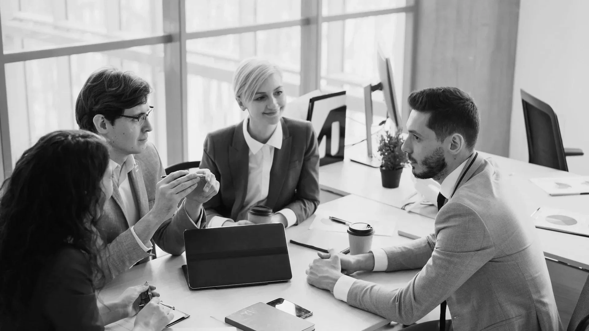 Five professionals in business attire having a discussion in a modern office with large windows, laptops, notebooks, and coffee cups on the table.