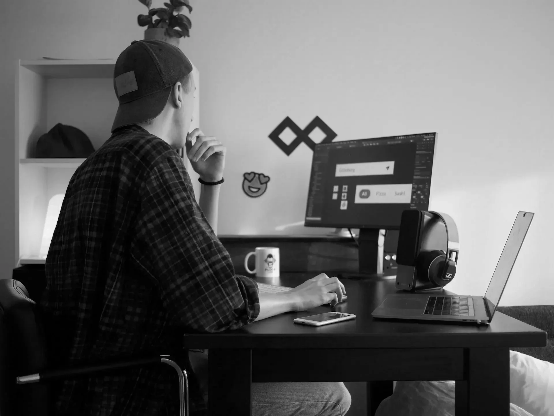 A young man wearing a baseball cap and a plaid shirt sitting at a desk in front of a computer monitor with design software open. The desk has a mug with face illustrations, a smartphone, and a pair of headphones. The background has a shelf with a plant and decorative stickers on the wall.