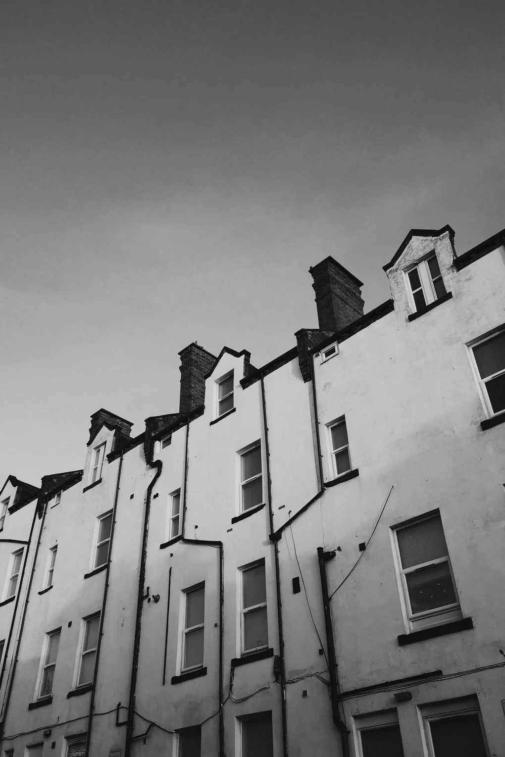 Black and white photo of the upper floors of old residential buildings with chimneys, windows, and doorways visible against a pale sky.