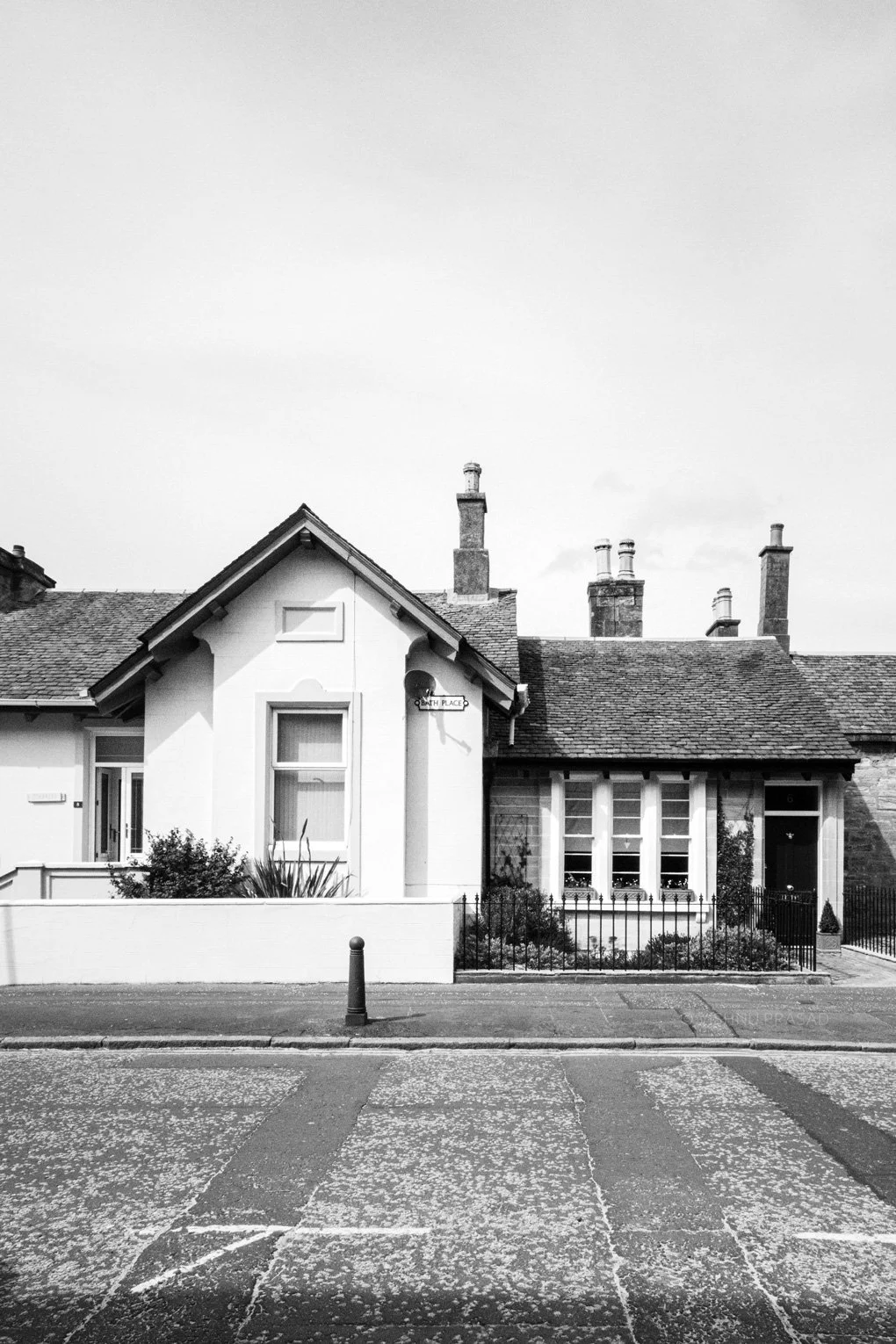 Black and white photo of residential houses with a sidewalk and street in front, featuring a mix of architectural styles, chimneys, and a small garden area.