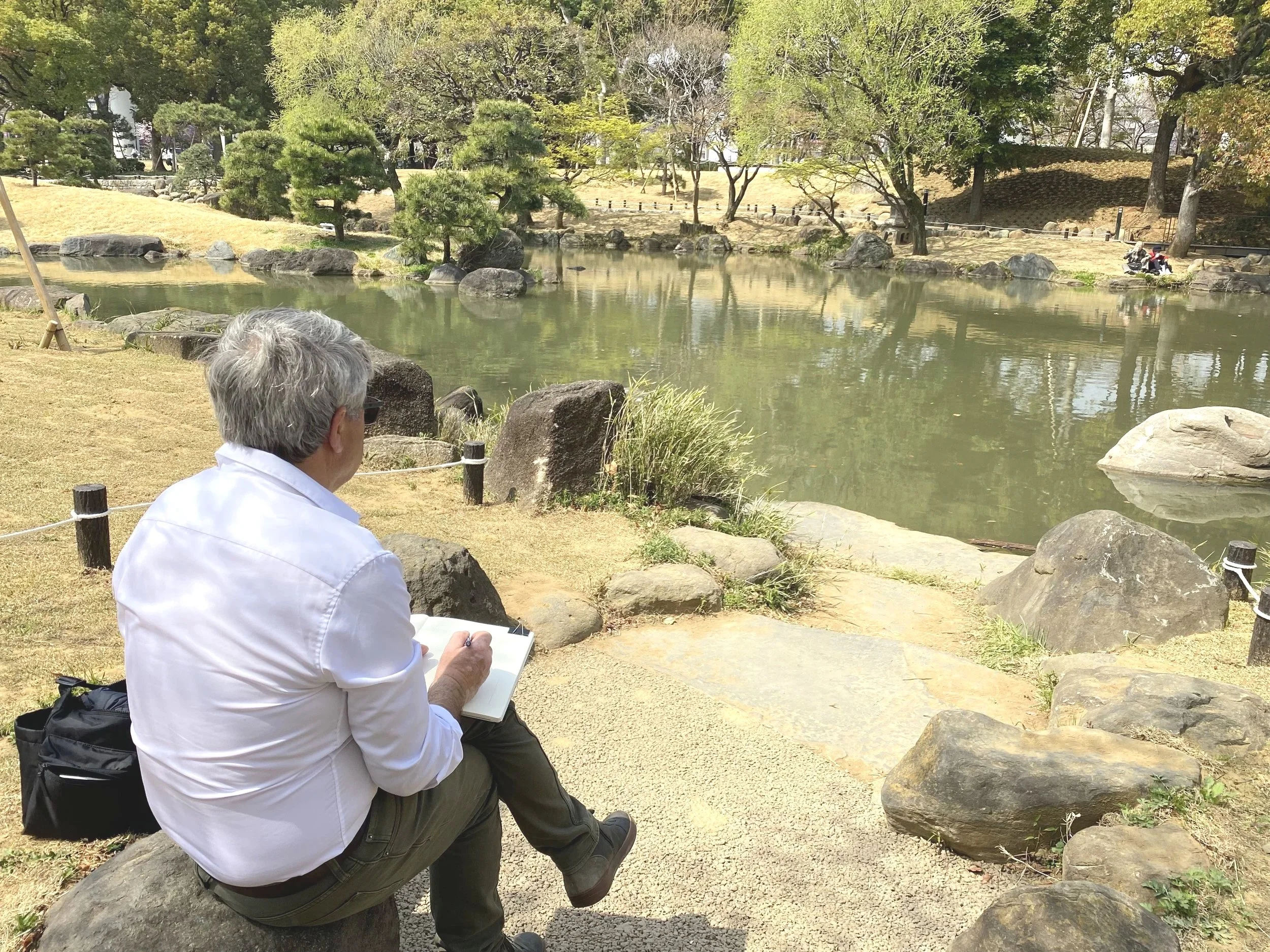 Une personne assise au sol dans un jardin japonais près d'un étang, en train de dessiner sur un carnet, avec des arbres et des rochers près de l'étang.