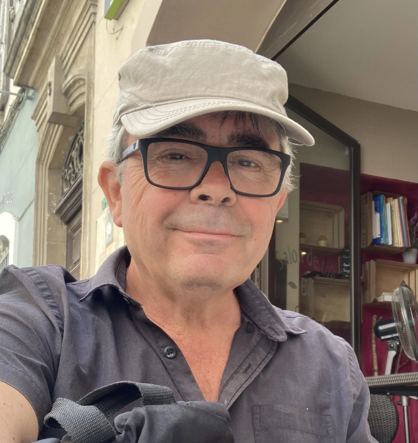 Un homme souriant portant une casquette beige, des lunettes noires et une chemise grise dans un magasin avec des étagères pleines de livres.