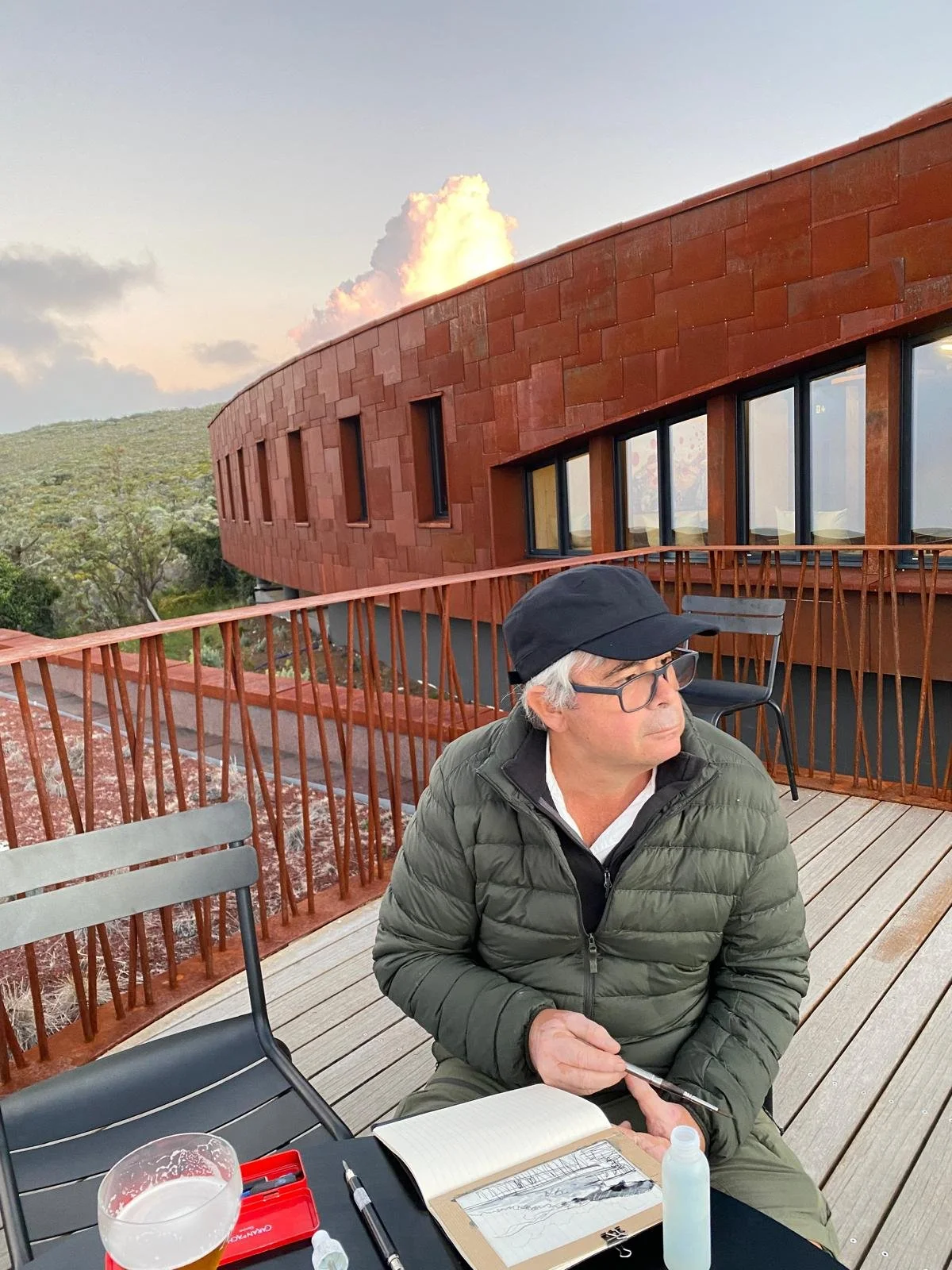 Un homme âgé portant une casquette noire, lunettes et un manteau vert assis sur une terrasse en bois avec une vue sur la nature. Il y a une table avec un carnet, un stylo, un verre de bière, un spray désinfectant, un masque et d'autres objets.