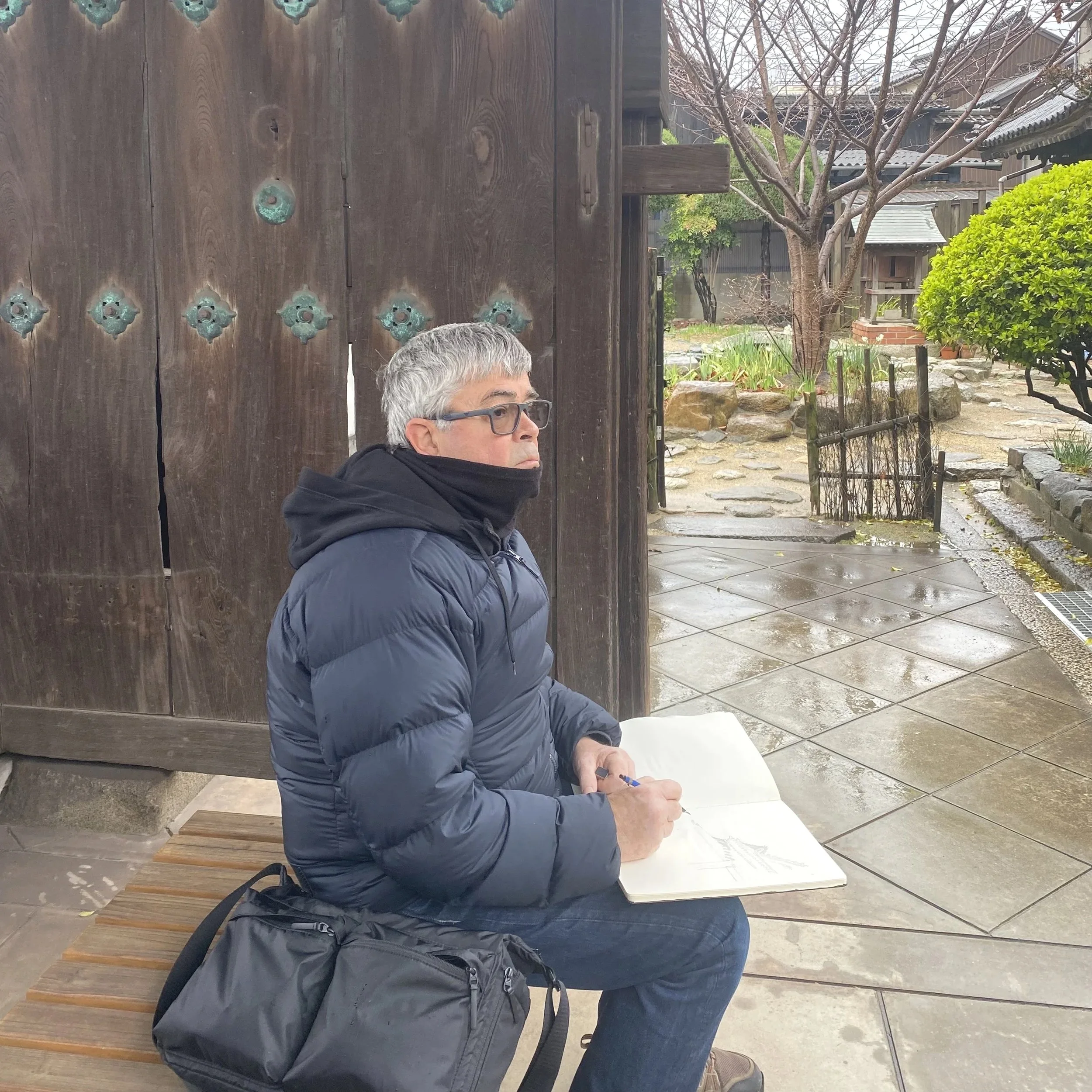L'artiste , assis sur un banc en bois, dessine sur un carnet blanc, en extérieur avec un arbre nu et une maison en arrière-plan.