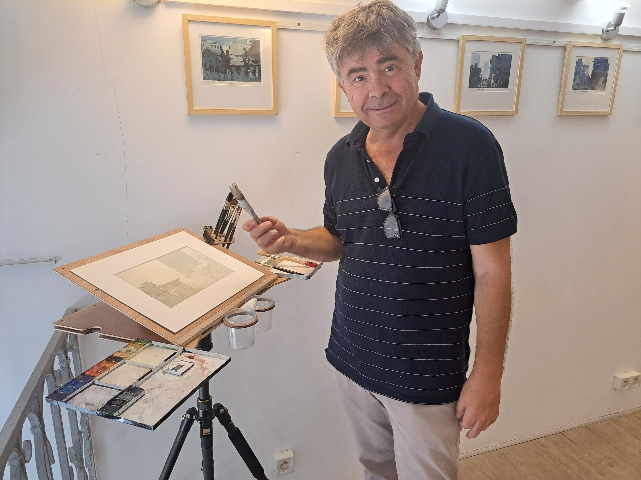 Un homme souriant, portant une chemise rayée noire et blanche, se tient dans un atelier d'art avec des cadres et des peintures suspendus au mur derrière lui. Il tient un pinceau dans sa main.