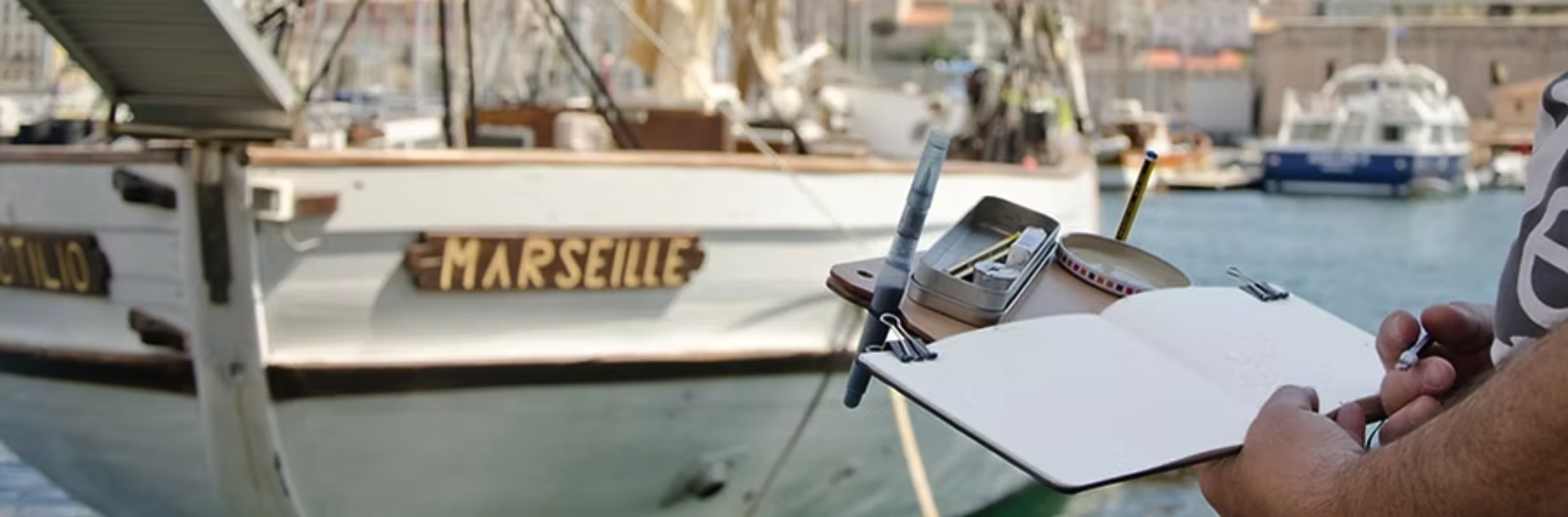 Person prenant des notes sur un carnet avec un bateau nommé « Marseille » en arrière-plan dans un port.