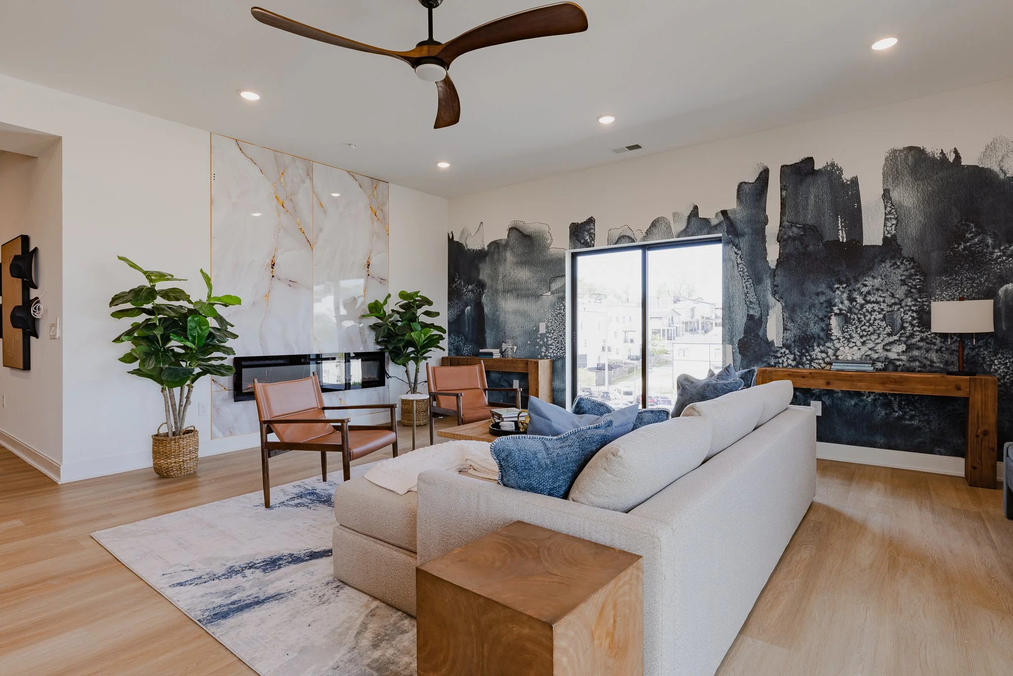 Modern living room with a beige sofa, wooden side table, blue armchair, abstract black and gray wall art, large window, ceiling fan, and houseplants.