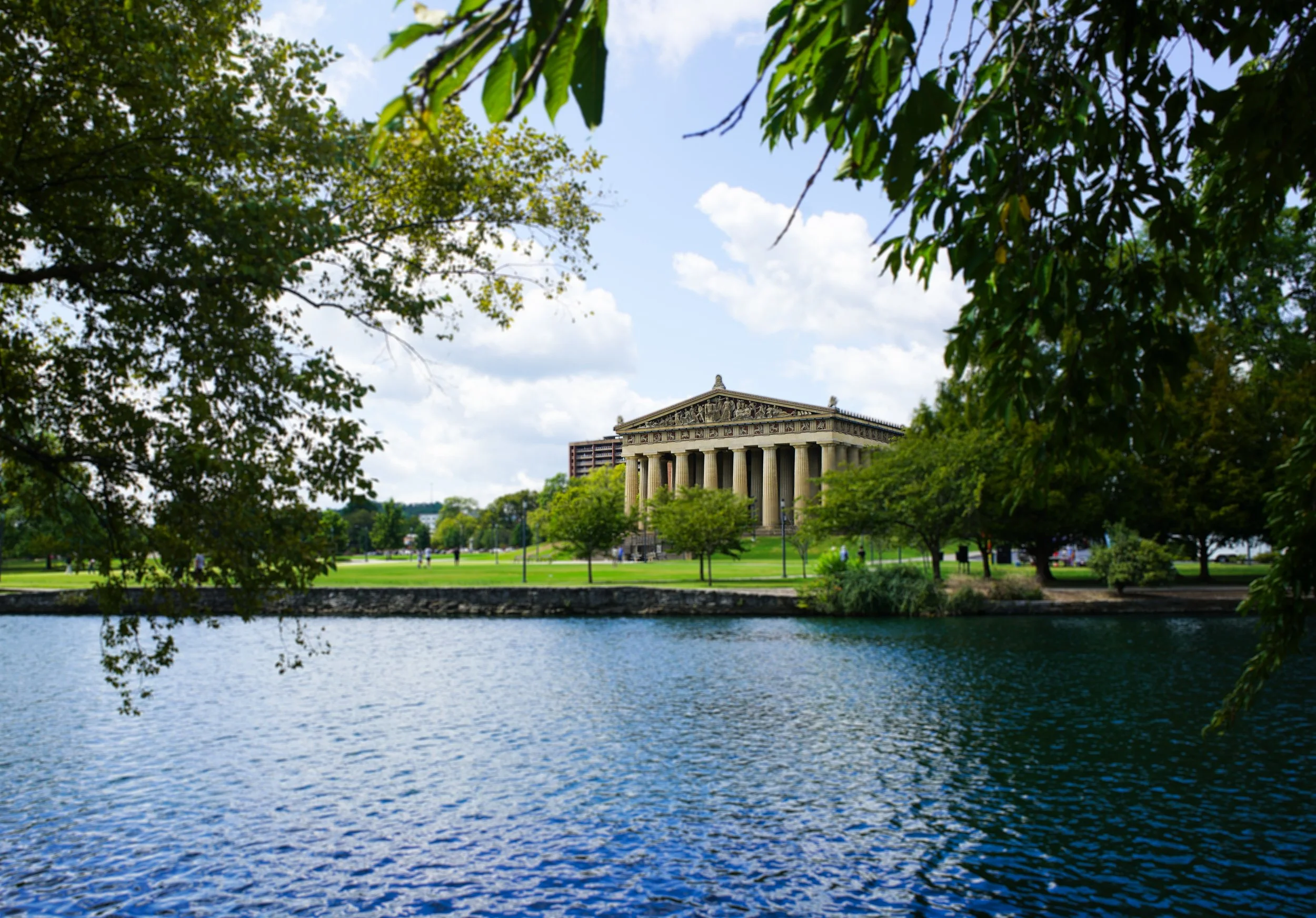 A view of a river with a historic Greek-style building with columns in the background, surrounded by green trees and a grassy park, under a partly cloudy sky.