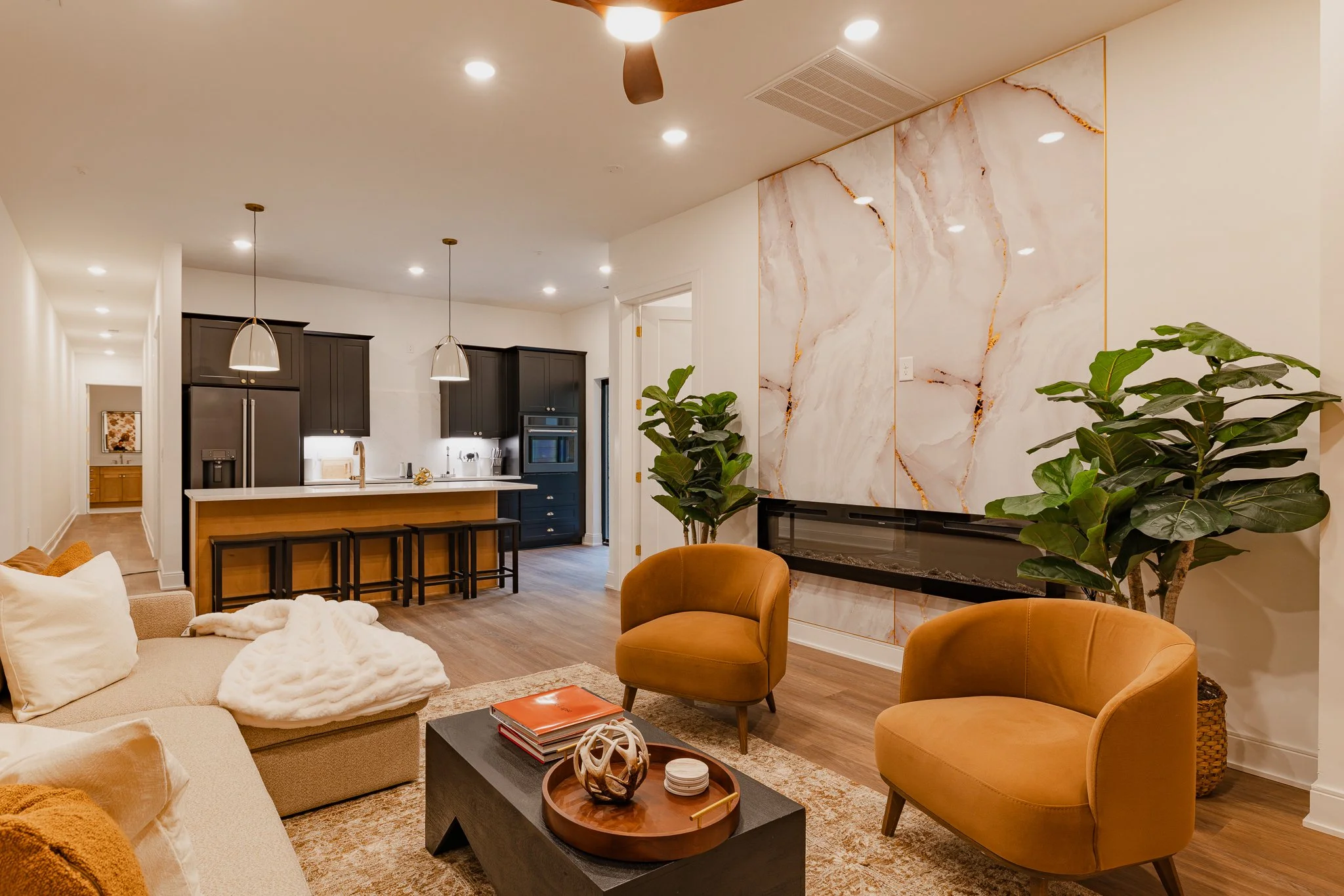 A modern living room and kitchen area with warm lighting, beige and brown furniture, a large marble style accent wall with potted plants, and pendant lights over a kitchen island with black stools.
