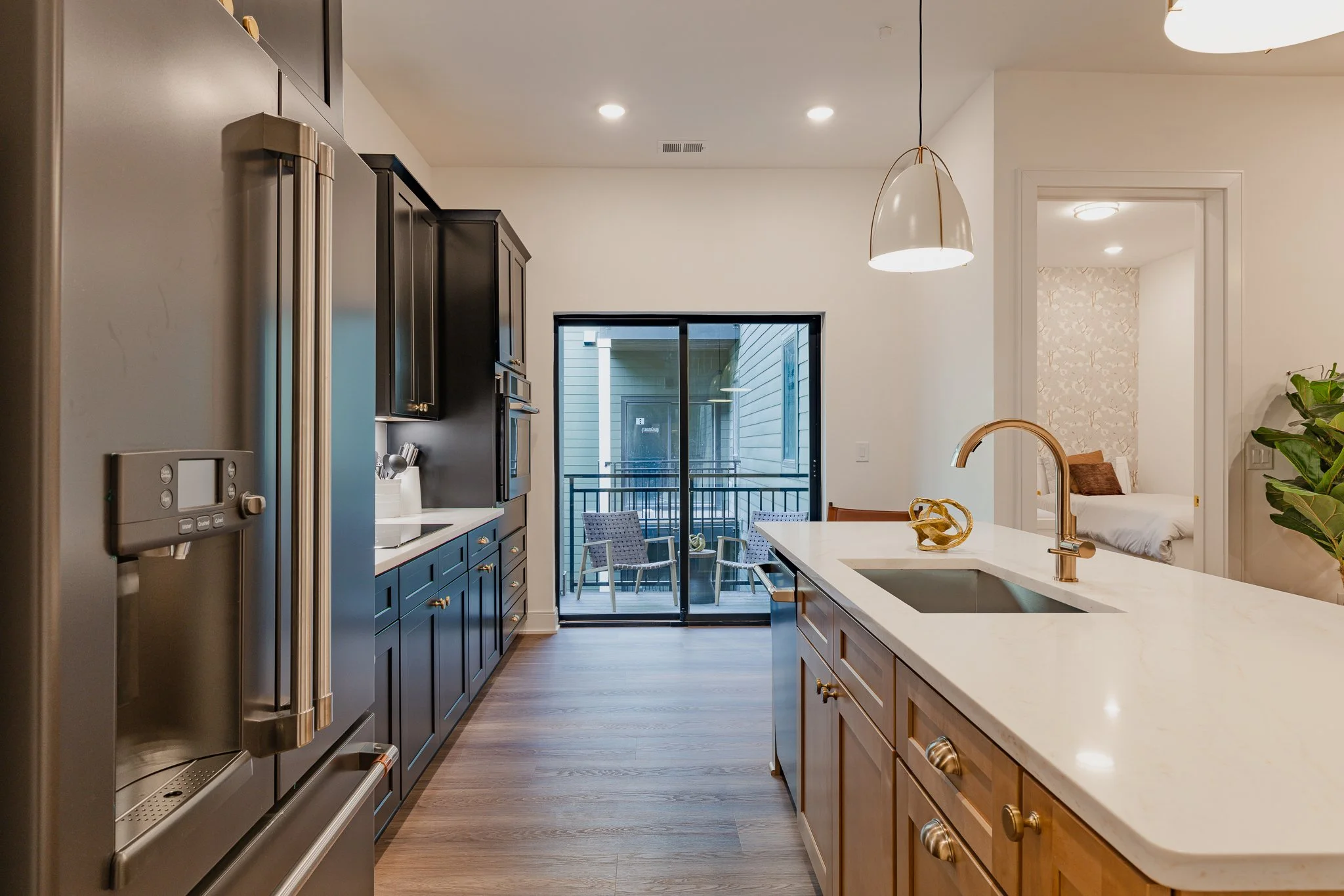 Modern kitchen with dark cabinets, stainless steel appliances, white marble countertops, and a view of a balcony with outdoor furniture. A bedroom with a bed and floral wallpaper is visible through an open doorway.