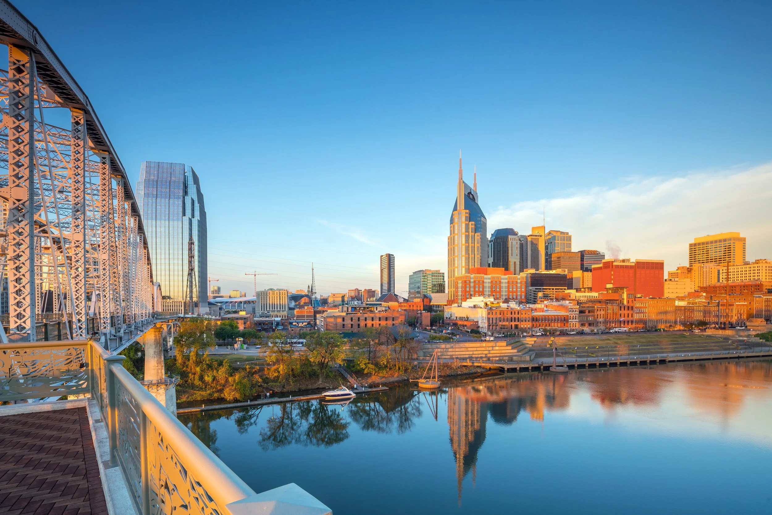 City skyline at sunset with a bridge over a river, featuring modern high-rise buildings and reflections in the water.