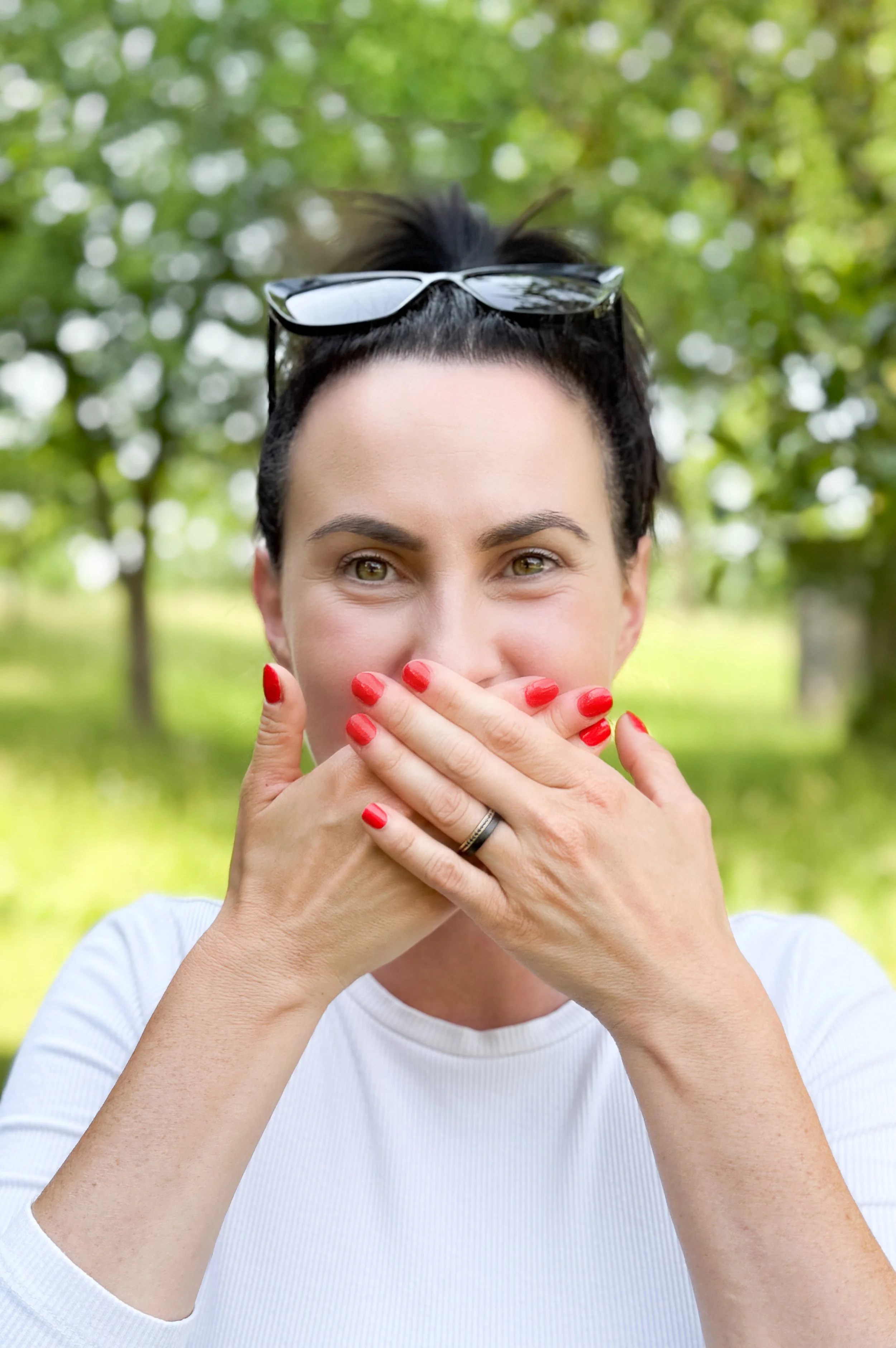 Eine Frau mit kurzen dunklen Haaren, die eine Brille auf dem Kopf trägt, bedeckt ihren Mund mit beiden Händen, die rote Nagellack haben, vor einem grünen, unscharfen Hintergrund.