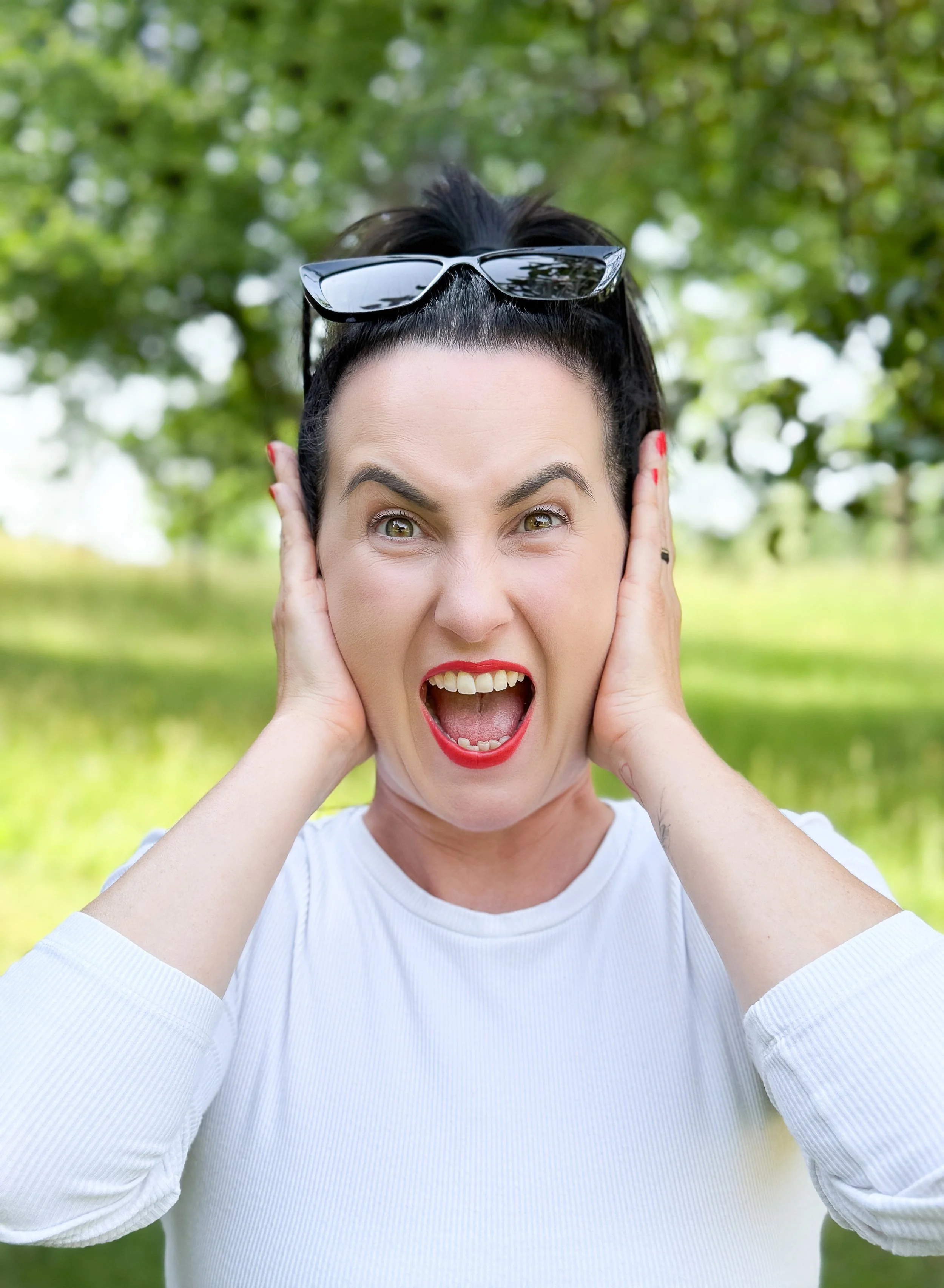 Eine Frau mit dunklem Haar trägt Sonnenbrille auf dem Kopf, hat rote Lippen, ist draußen in der Natur, wirkt überrascht oder erschrocken und hält sich die Ohren mit den Händen.