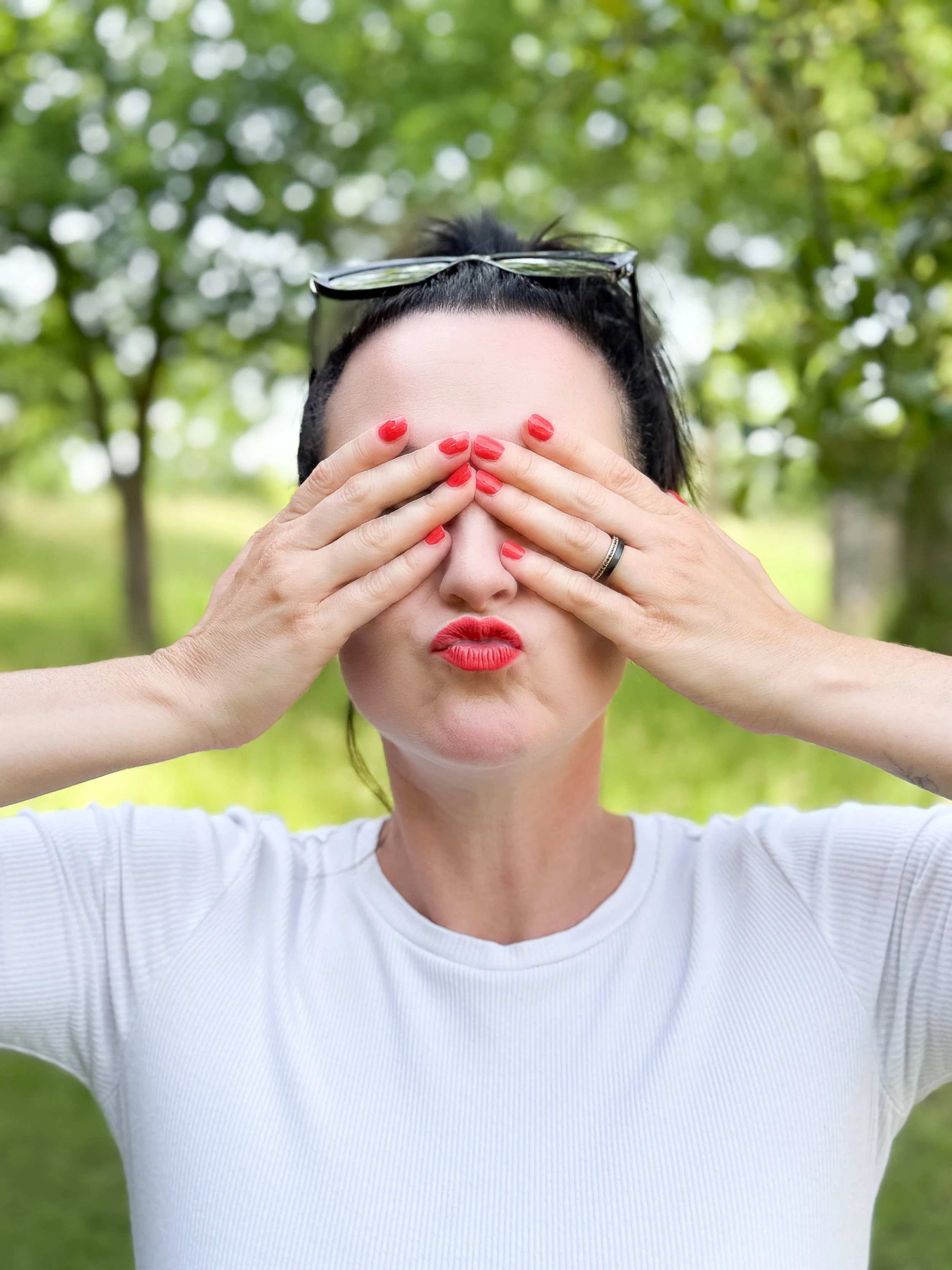 Eine Frau mit roten Lippen und roten Nägeln, die ihre Hände vor das Gesicht hält. Sie trägt eine weiße Oberbekleidung, hat schwarze Haare und eine Sonnenbrille auf dem Kopf. Der Hintergrund besteht aus grünen Bäumen im Freien.