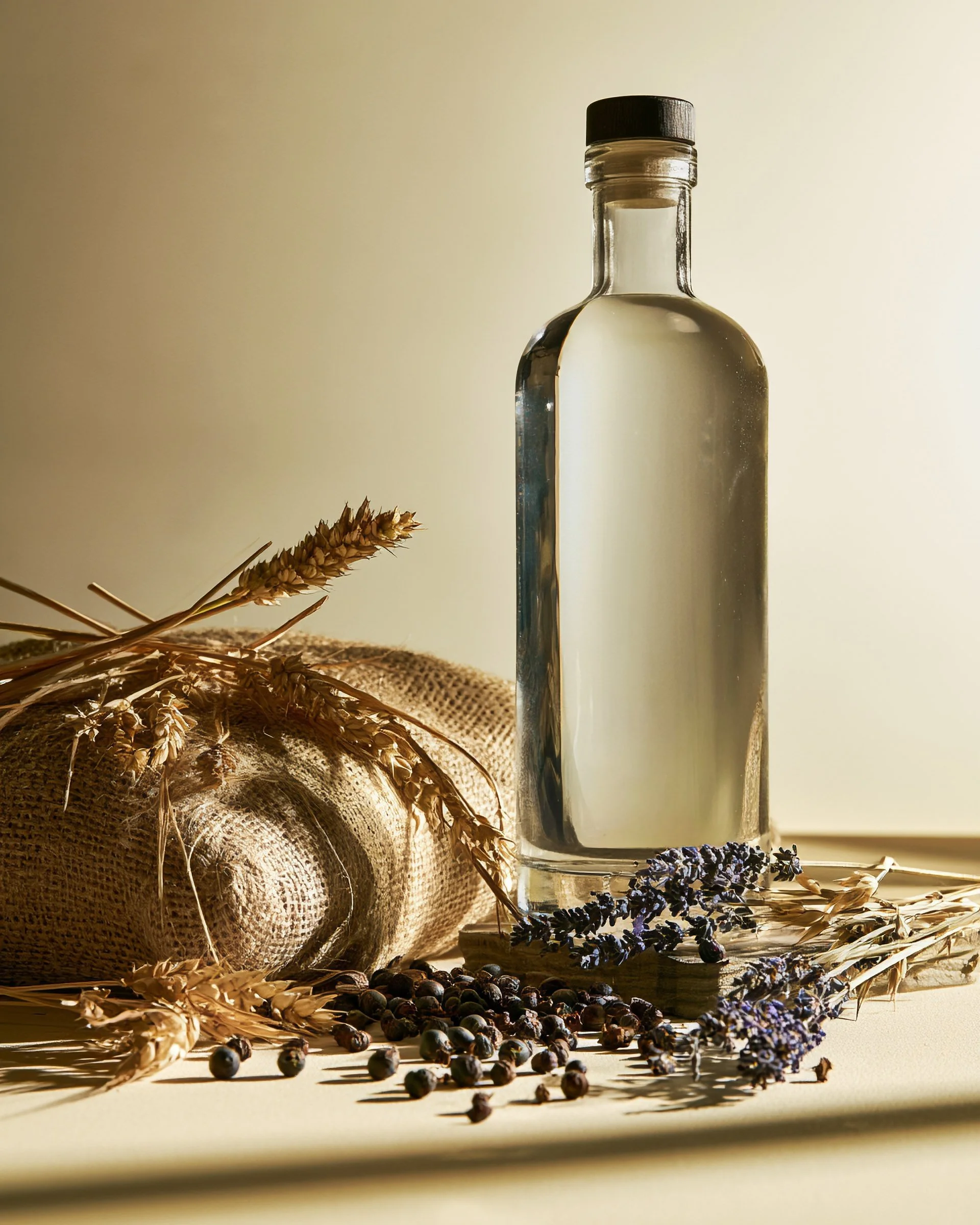 A glass bottle filled with a clear liquid, placed on a surface surrounded by dried lavender sprigs, wheat stalks, and scattered dark berries, with a soft light creating shadows and highlights.