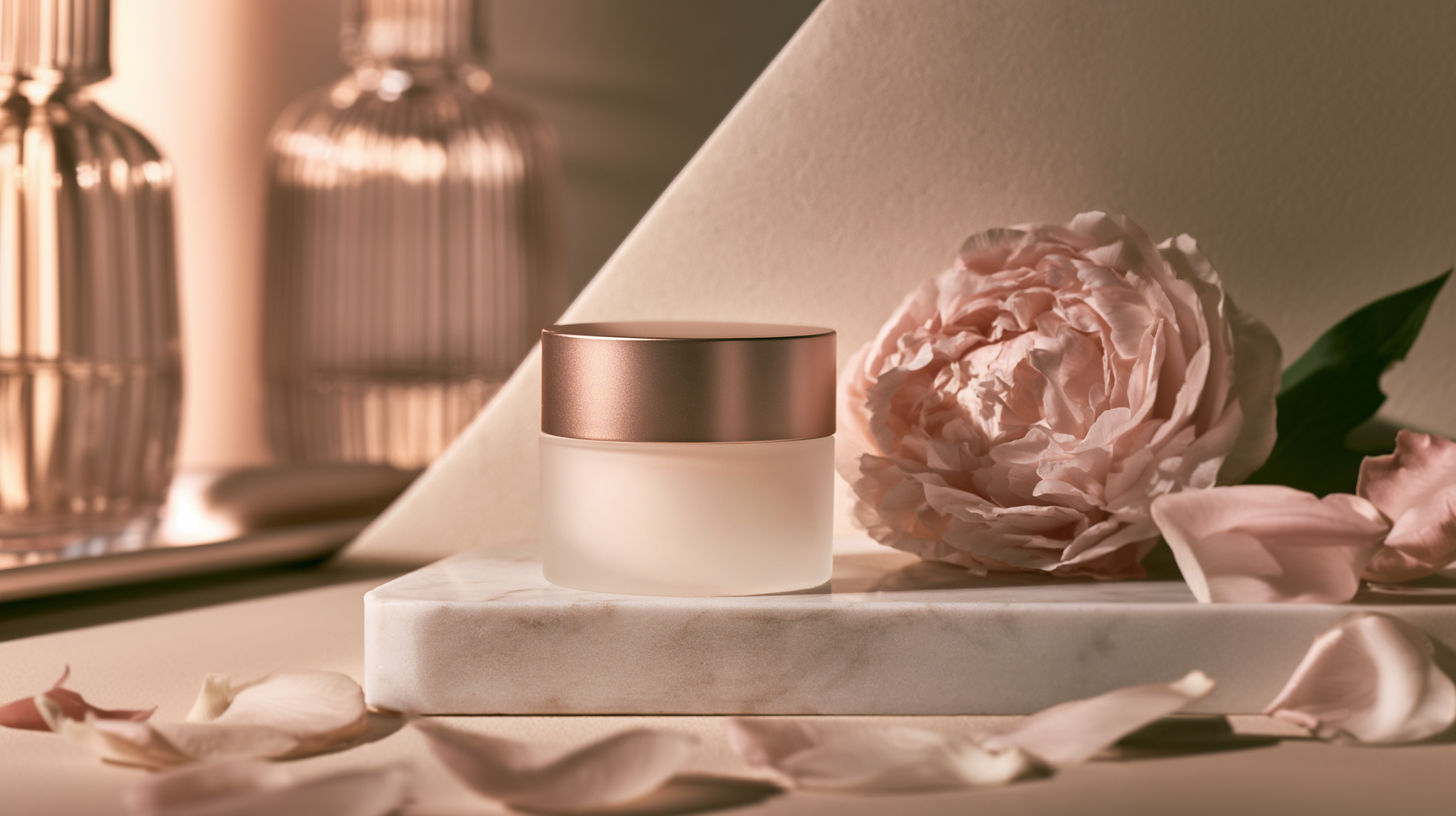 A white and rose gold cosmetic jar placed on a white marble surface, surrounded by a pink peony flower and petals, with a blurred background of decorative glass bottles.