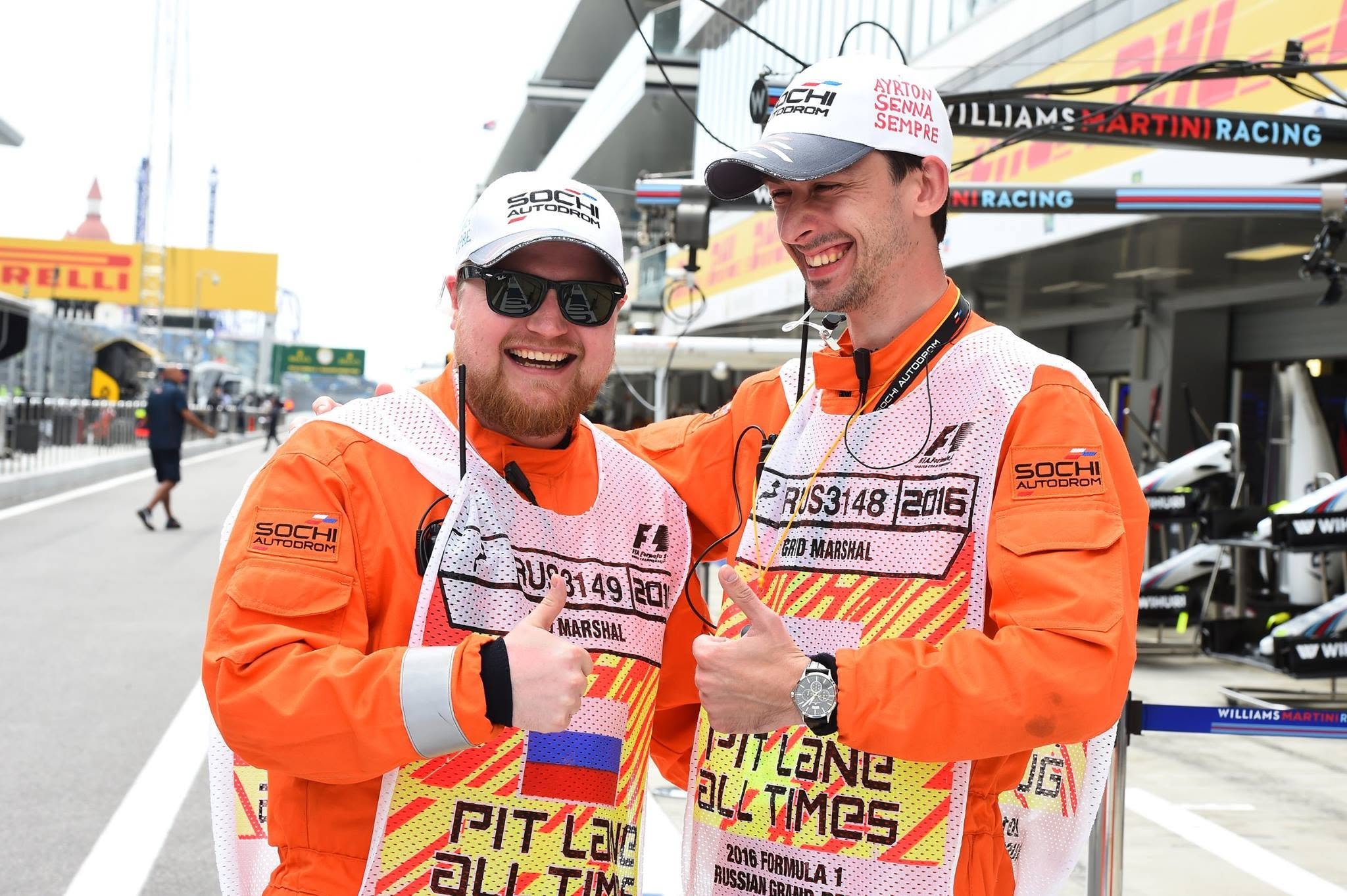 Two race marshals in orange uniforms standing in pit lane during a motorsports event.