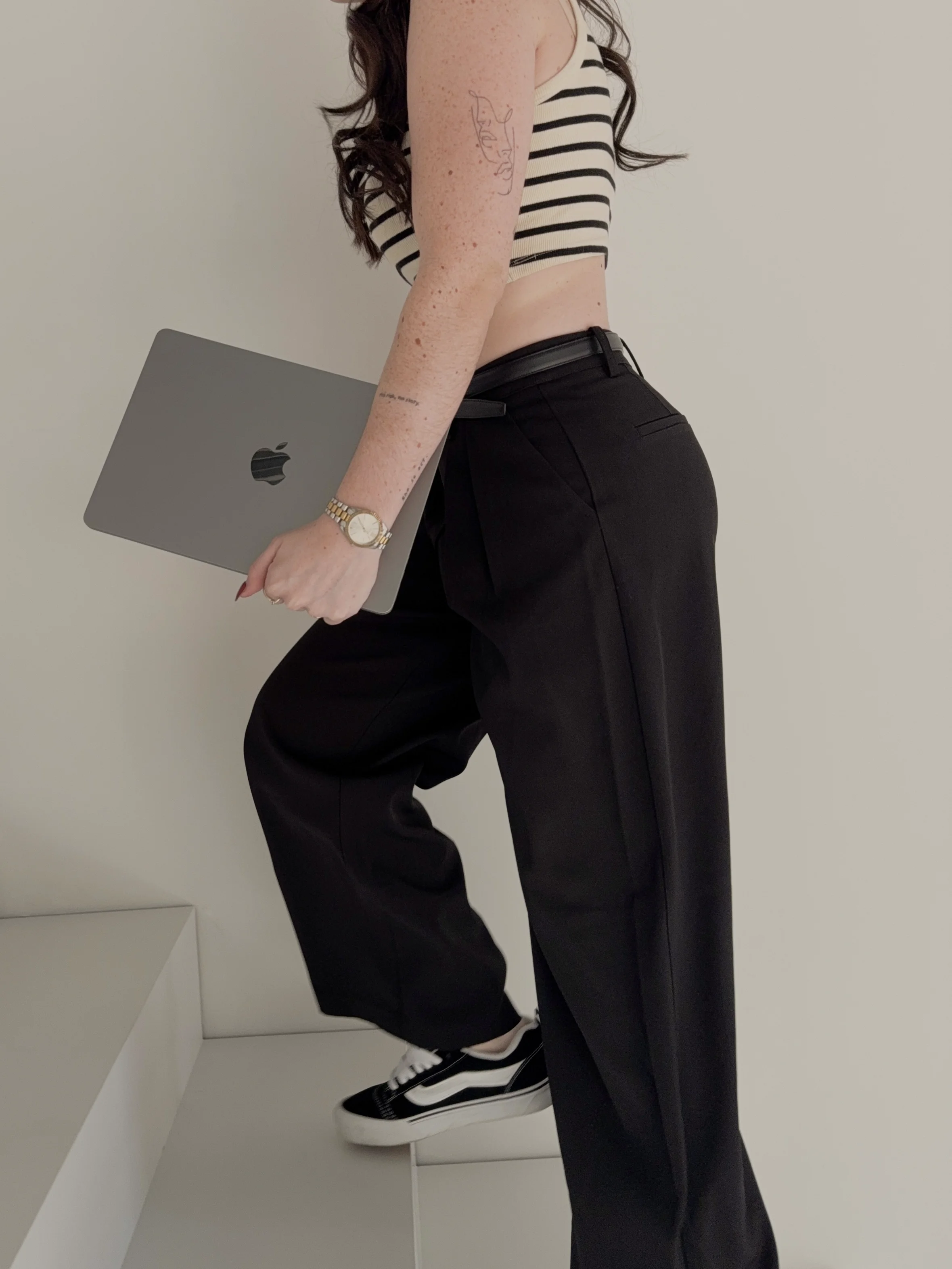 Britt Breemen social media manager, in zwarte broek, zwart-wit gestreept t-shirt en sneakers, met een Apple MacBook in de hand, staand in een kamer met witte muren.