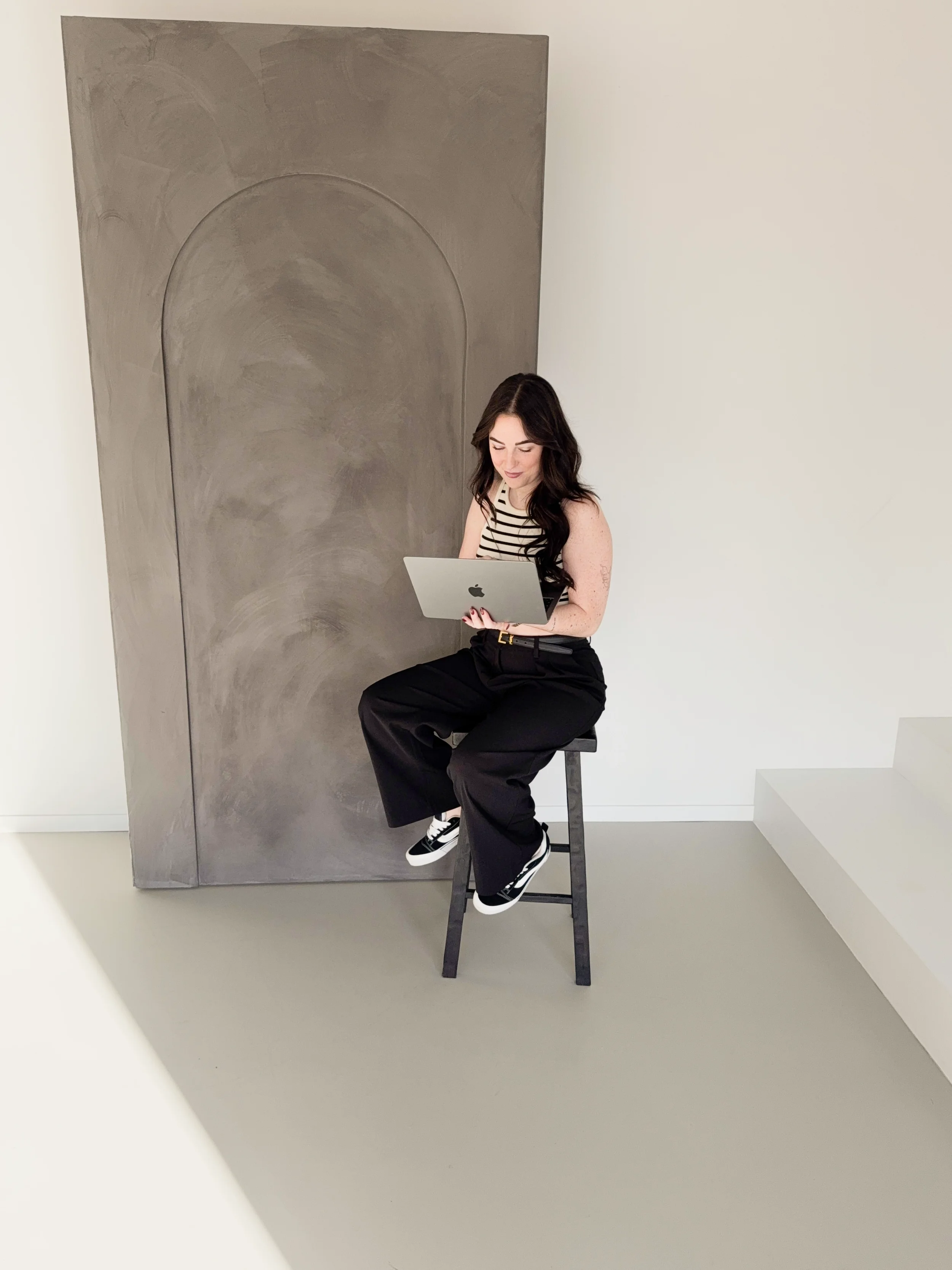 Britt Breemen social media manager, zit op een kruk, kijkt naar haar laptop, tegen een abstract muur met grijstinten.