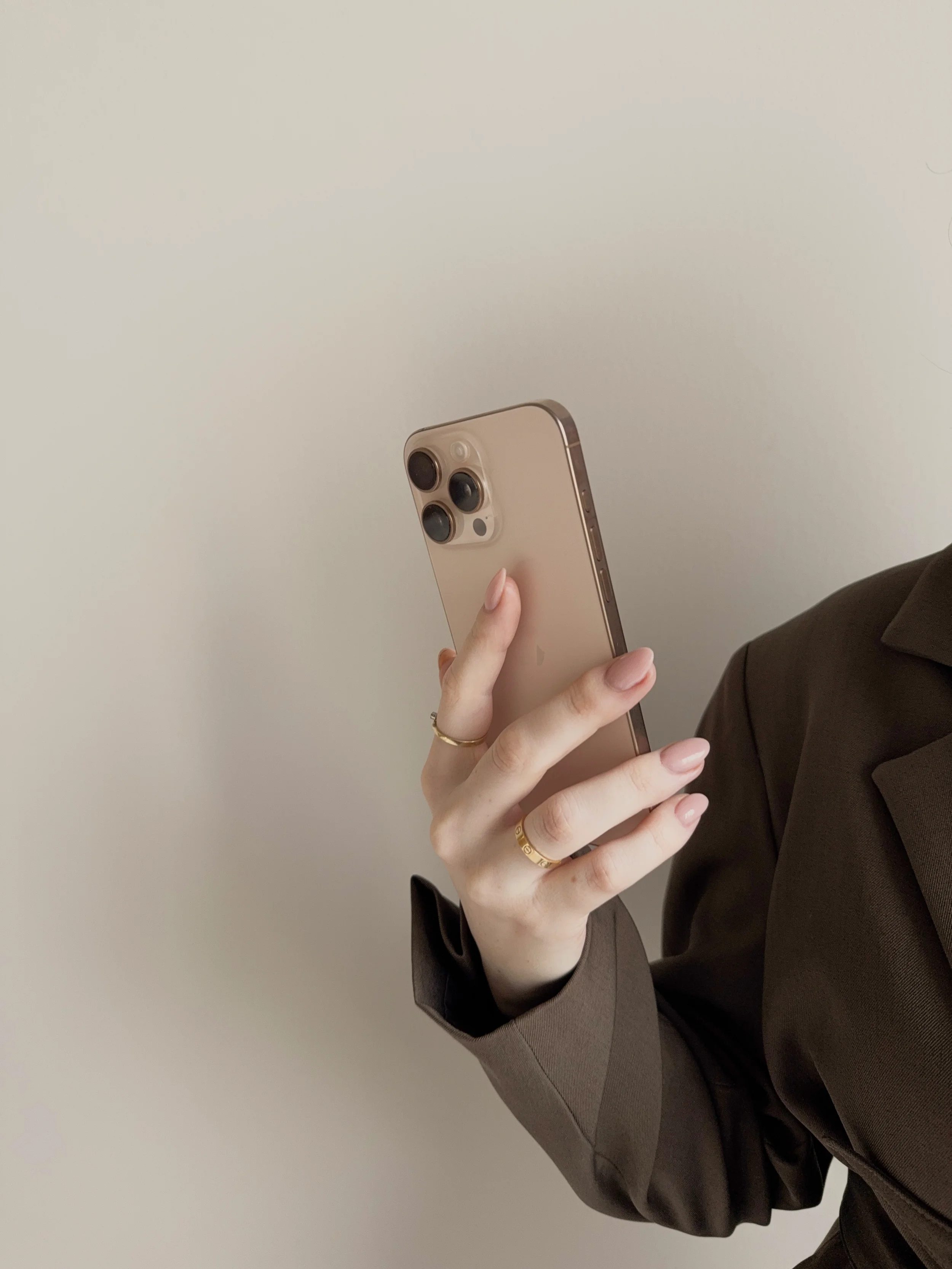 Britt Breemen social media manager, close-up van een hand met goudkleurige ring die een gouden iPhone vasthoudt, gekleed in een donkerbruin jasje, tegen een effen lichtgrijze achtergrond.