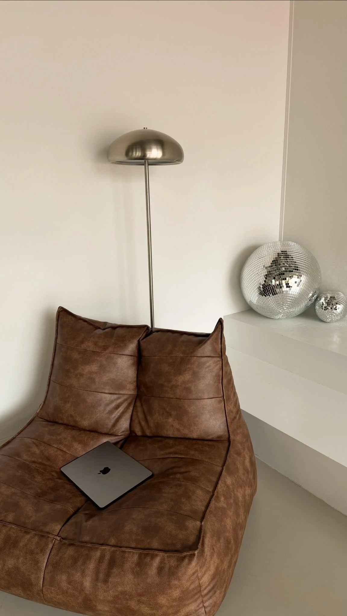 Een leren bank met een open laptop ernaast, een staande metalen lamp, en decoratieve disco ballen op een witte plank in een modern interieur.