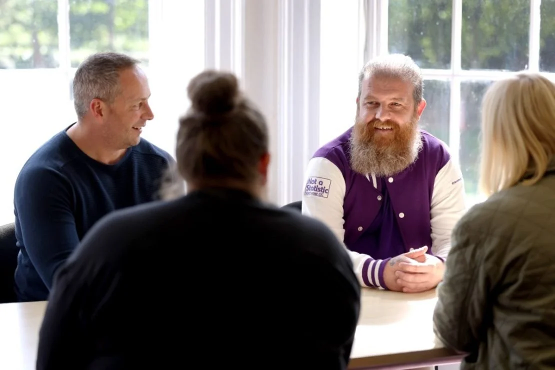Five people having a conversation around a table, with large windows in the background letting in natural light.