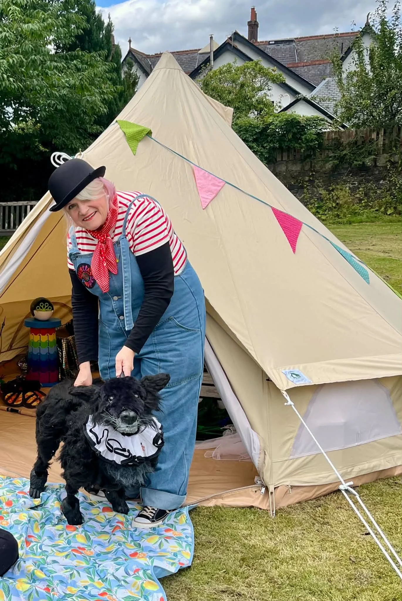 Myette Godwyn in a bowler hat and denim dungarees with a black dog puppet beside a bell tent.