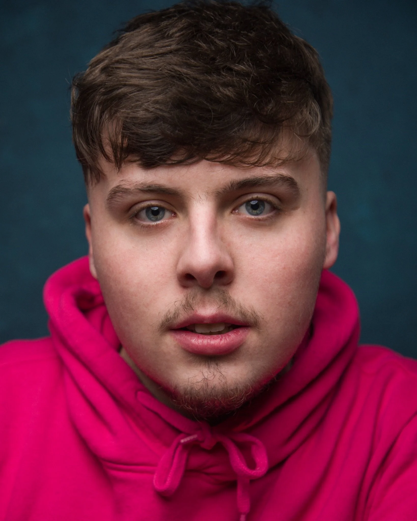 Headshot of actor Cieran Low in a bright pink hoodie against a dark blue background.