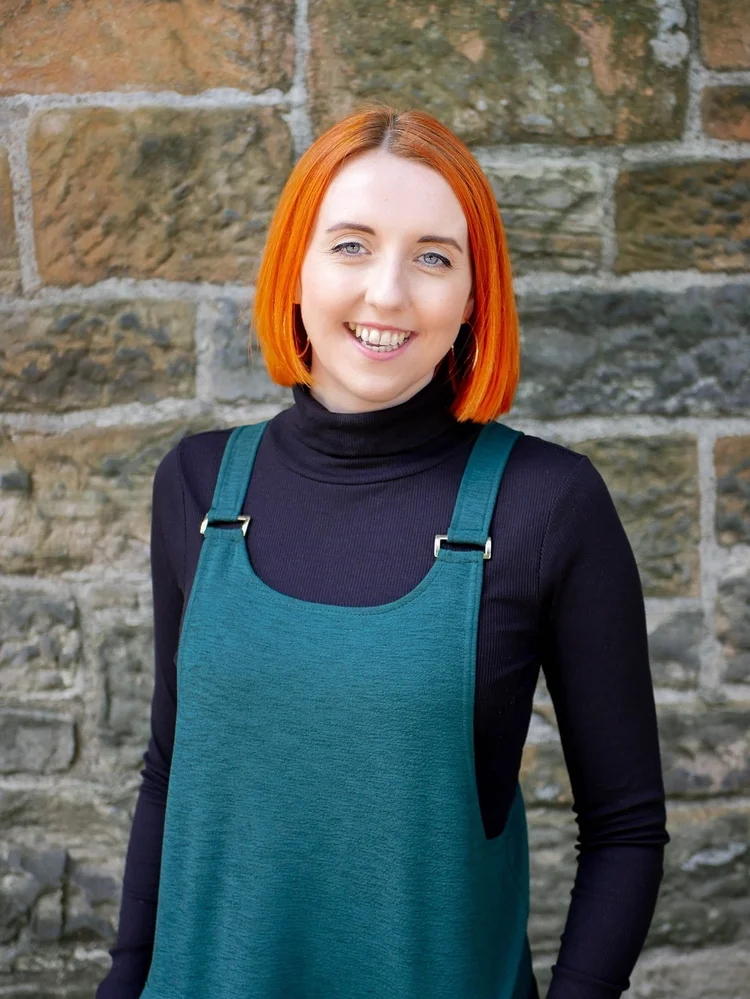 Portrait of director Sarah Marie Mooney smiling in a teal dress and black top against a stone wall.