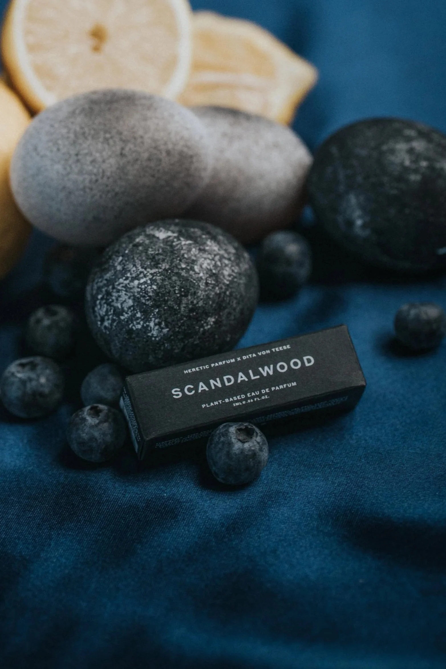 Close-up of a collection of black, gray, and white speckled stones, with a box labeled 'Scandalwood' perfume and sliced lemons in the background, all arranged on a dark blue fabric surface.