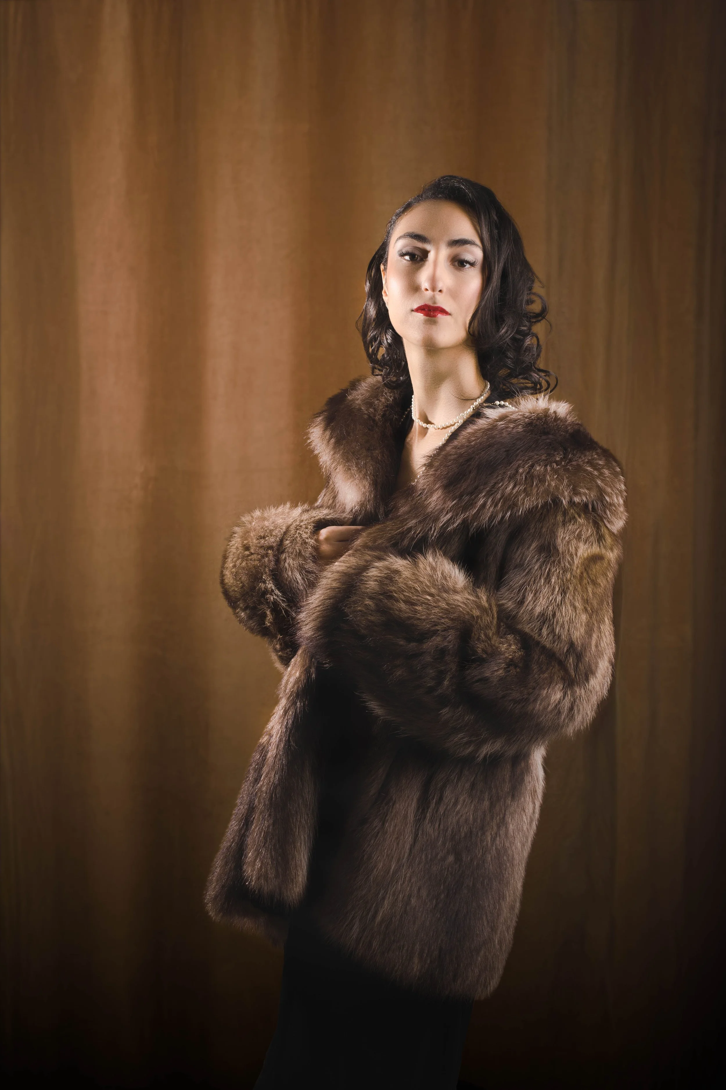 A woman with dark, curled hair, wearing a pearl necklace, red lipstick, and a luxurious brown fur coat, posing against a brown curtain background.