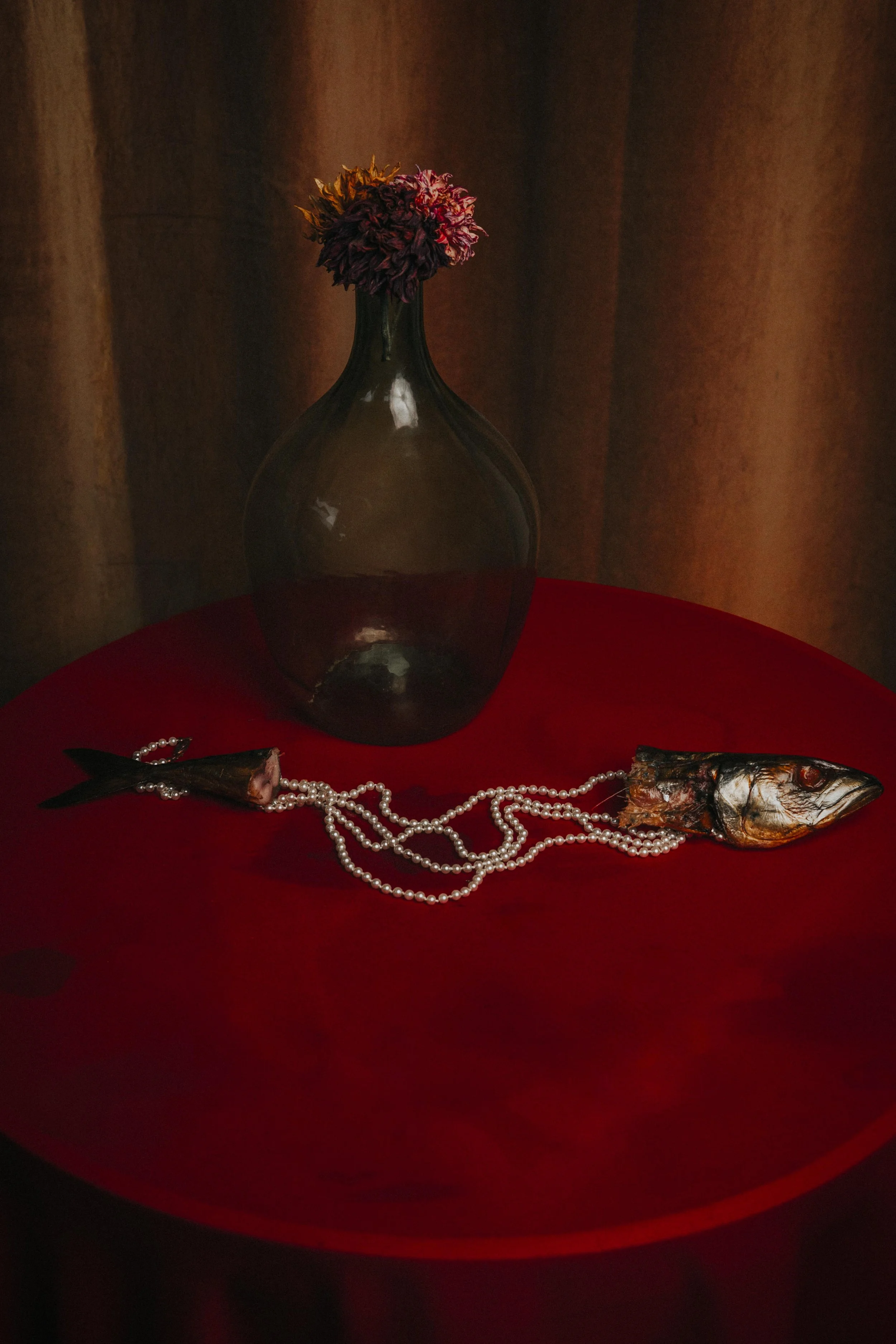 A dark, transparent glass vase with flowers on top, placed on a red cloth-covered surface. A fish wrapped in foil and a pearl necklace are also on the surface. The background features a brownish curtain.
