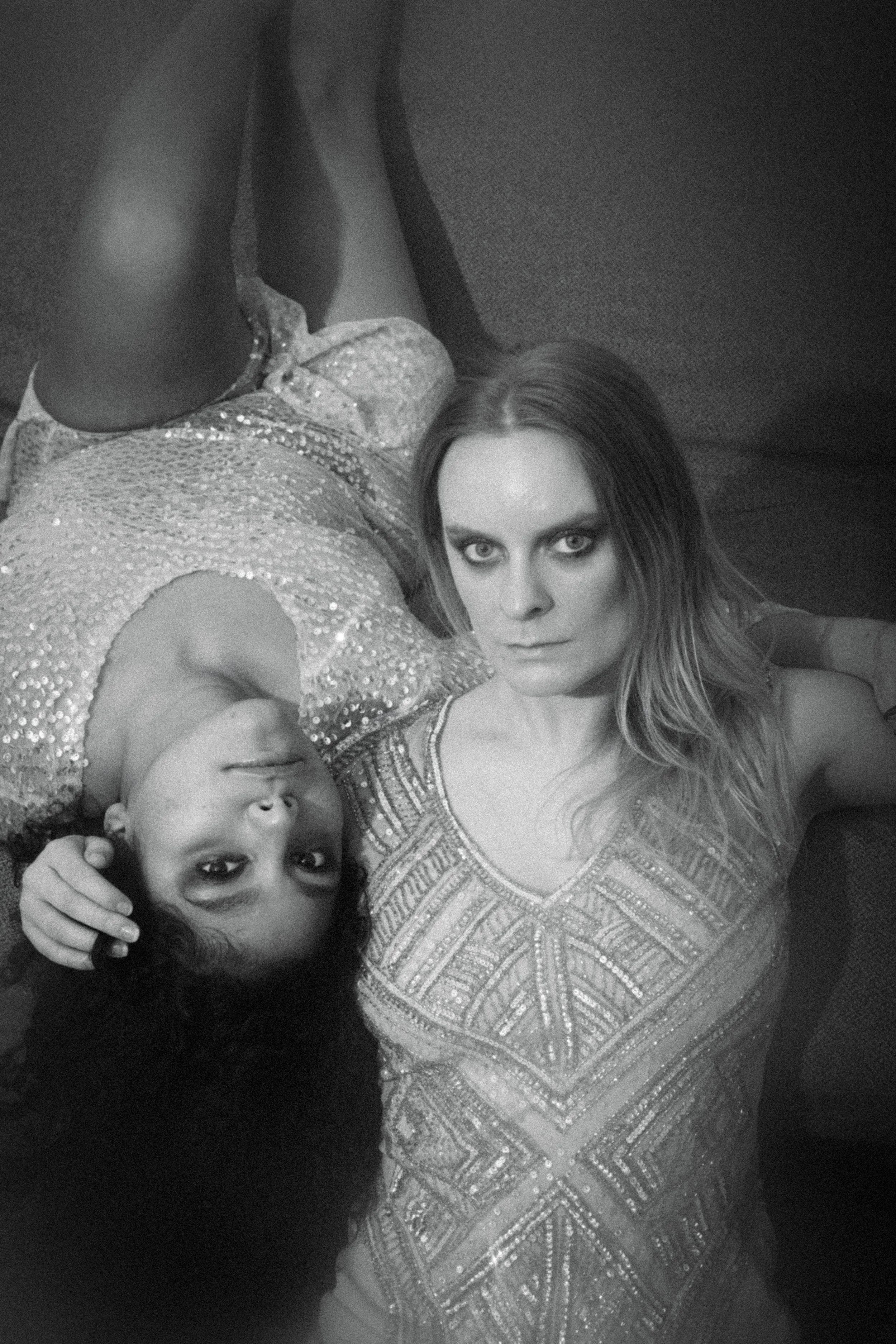 A woman and a young girl lying down together. The woman has long hair and wears a patterned top, looking directly at the camera. The girl has curly hair, resting her head near the woman's and looking at the camera, smiling.