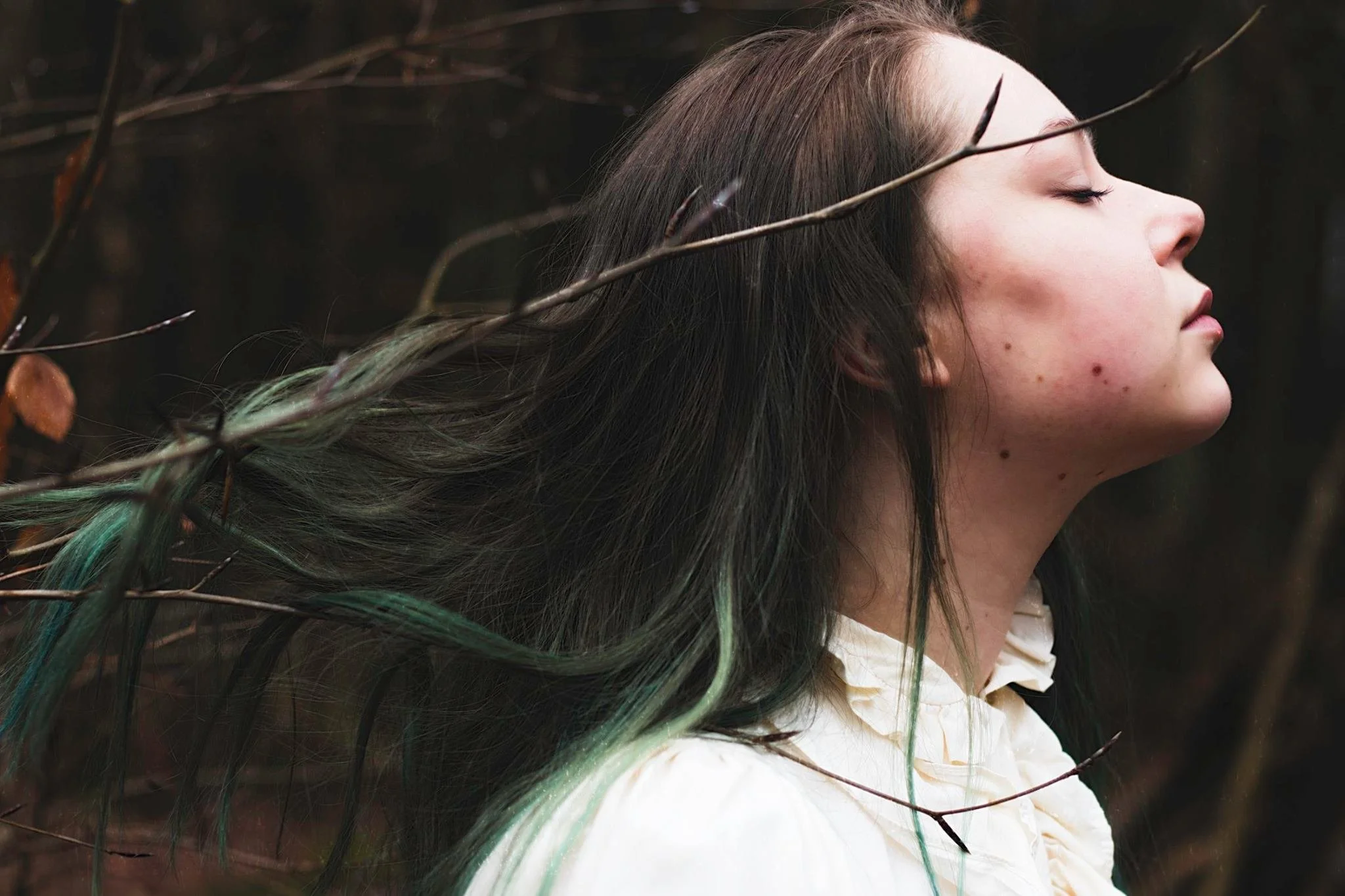 Profile of a woman with long brown hair with green tips, standing outdoors among bare branches, eyes closed, wearing a cream-colored blouse.