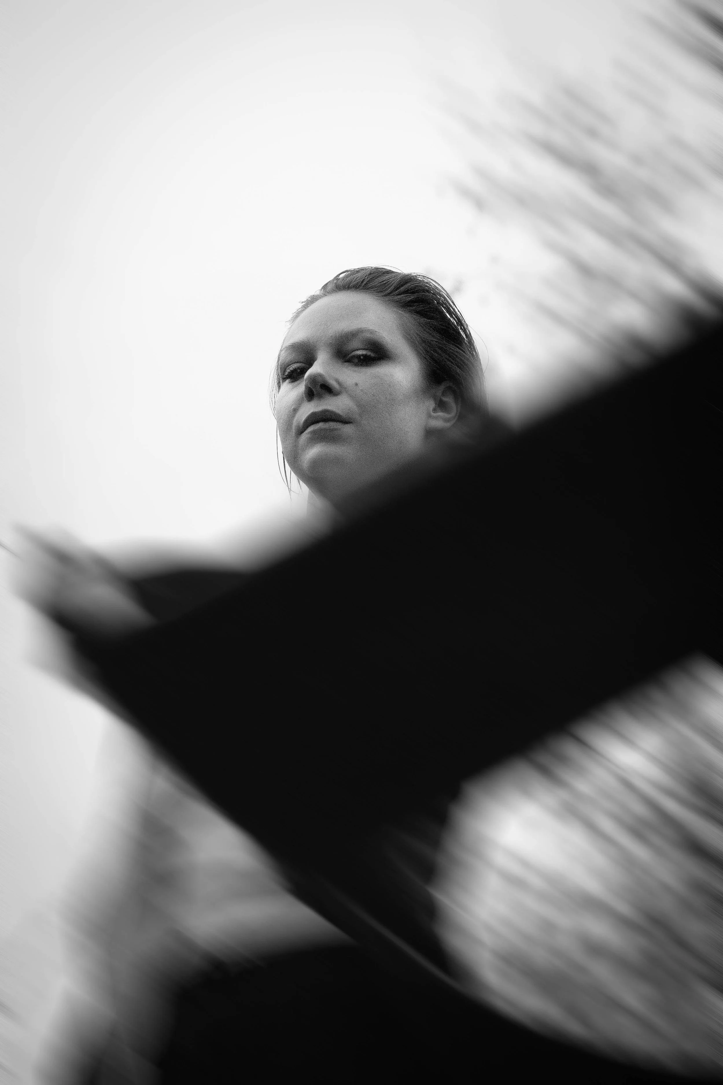 Black and white photograph of a woman with slicked-back hair, looking at the camera with an intense expression. The image is taken from a low angle, with blurred lines or streaks in the foreground.