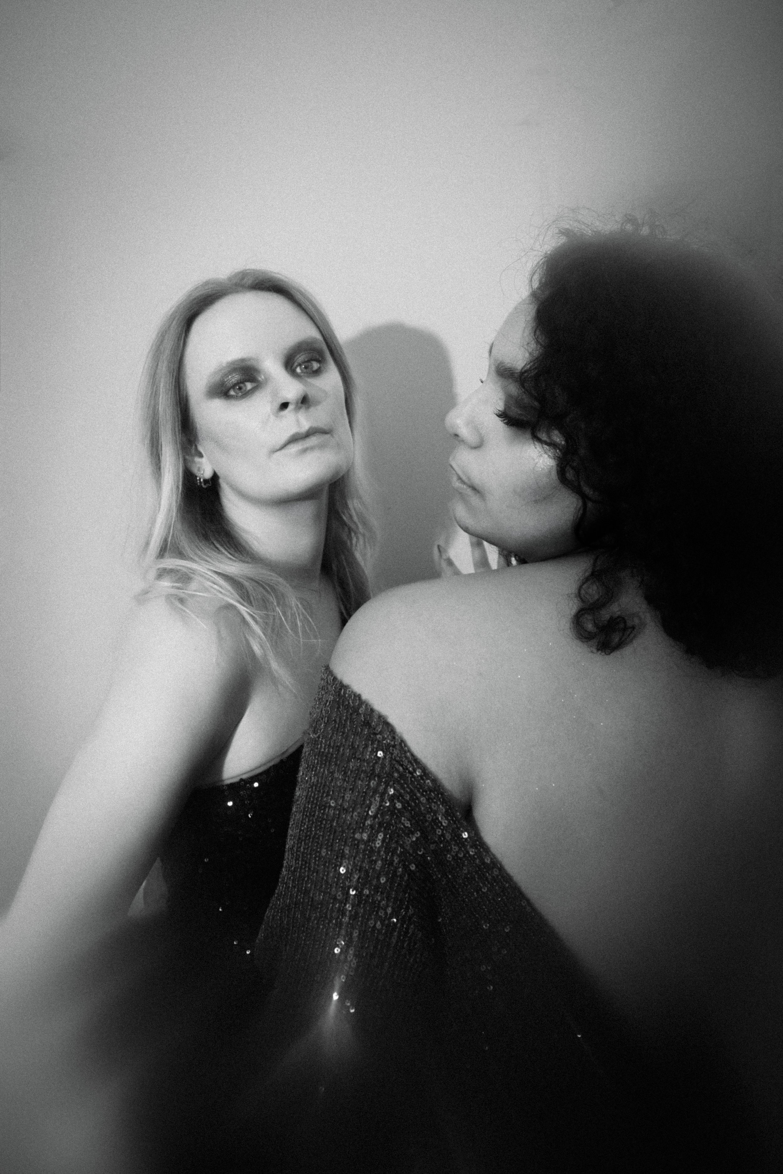 Two women in glamorous attire pose closely together for a black and white photo, one with light-colored hair and makeup, the other with dark, curly hair and adorned with sequined clothing.