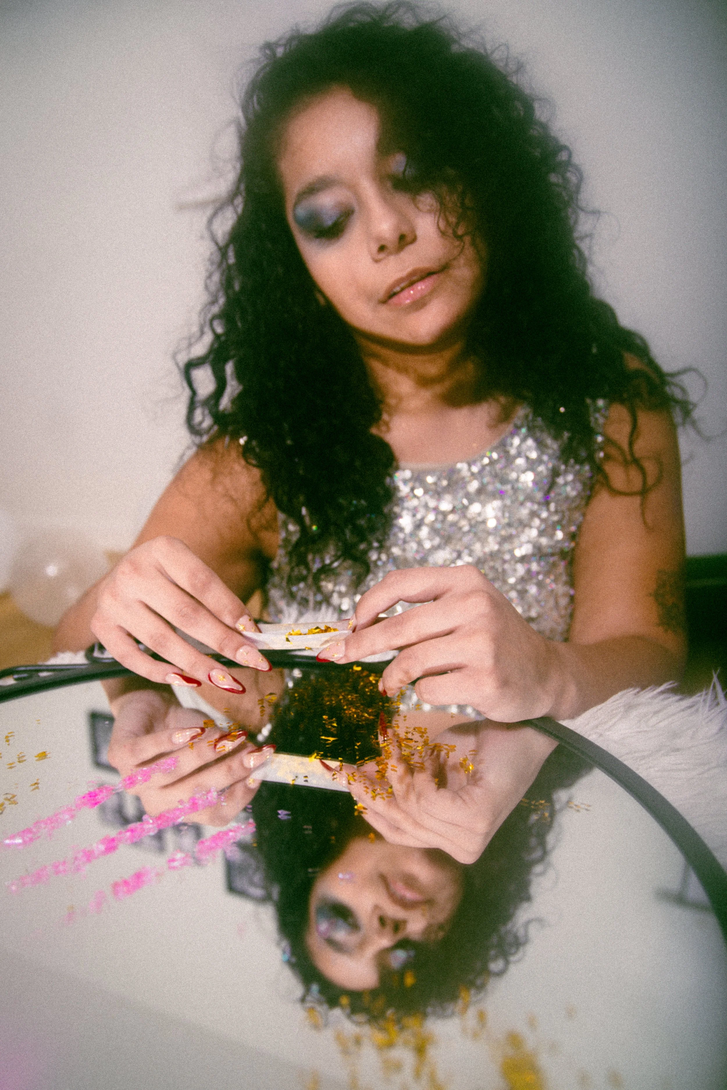 A woman with curly black hair, wearing a sparkly silver top, is sitting at a table with a mirror. She is looking down at her hands, which are holding a makeup palette. The table reflects her face and hands, and there are colorful glitter pieces scatt