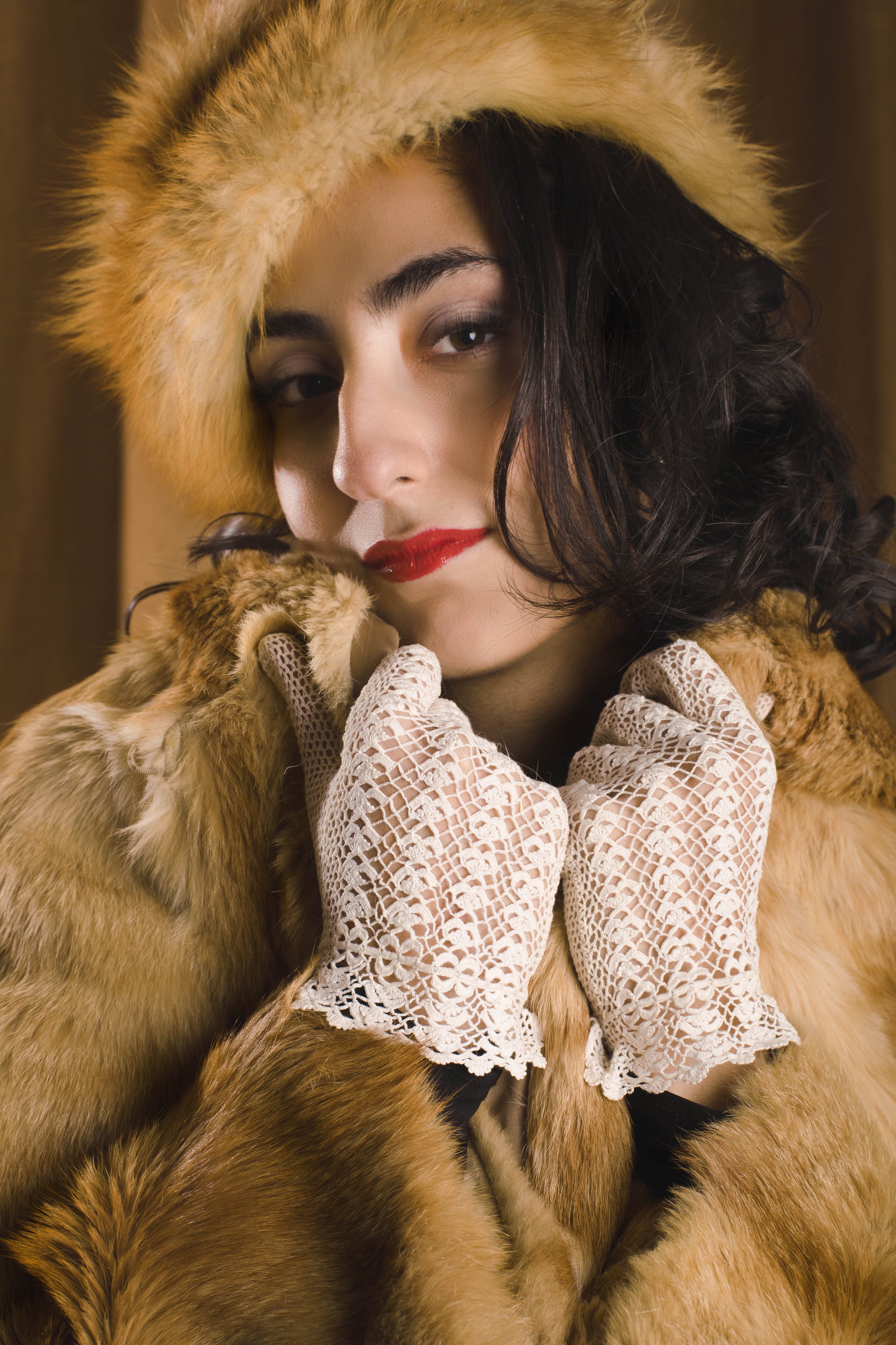 A woman with dark, curly hair wearing a large, fur-lined hooded coat and lace gloves, slightly smiling with red lipstick, looking into the camera.