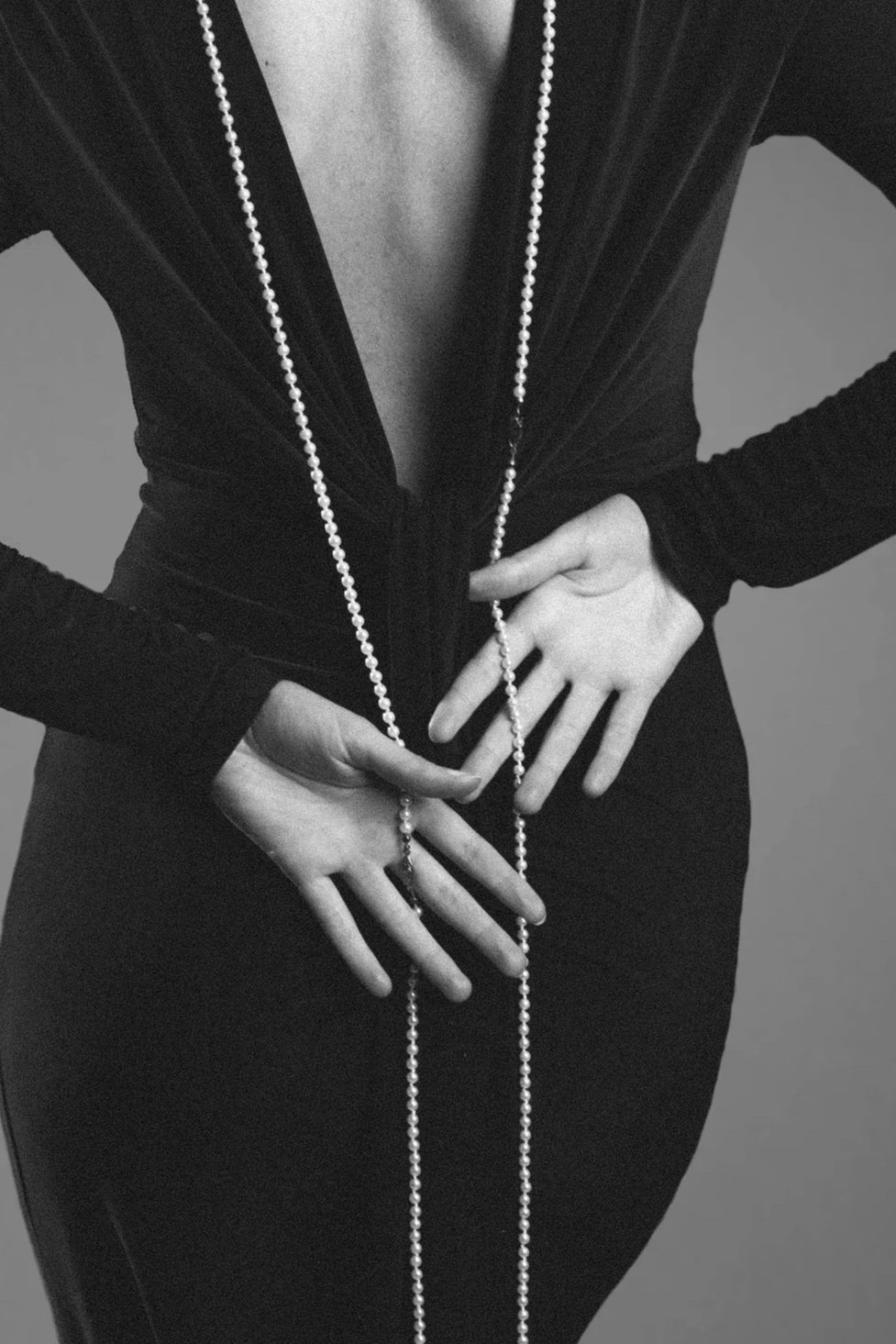 Black and white photo of a person wearing a black, long-sleeved dress with a deep V-neckline and a long pearl necklace. The person is holding the necklace with both hands, with one hand on the waist and the other near the hips, against a plain background.