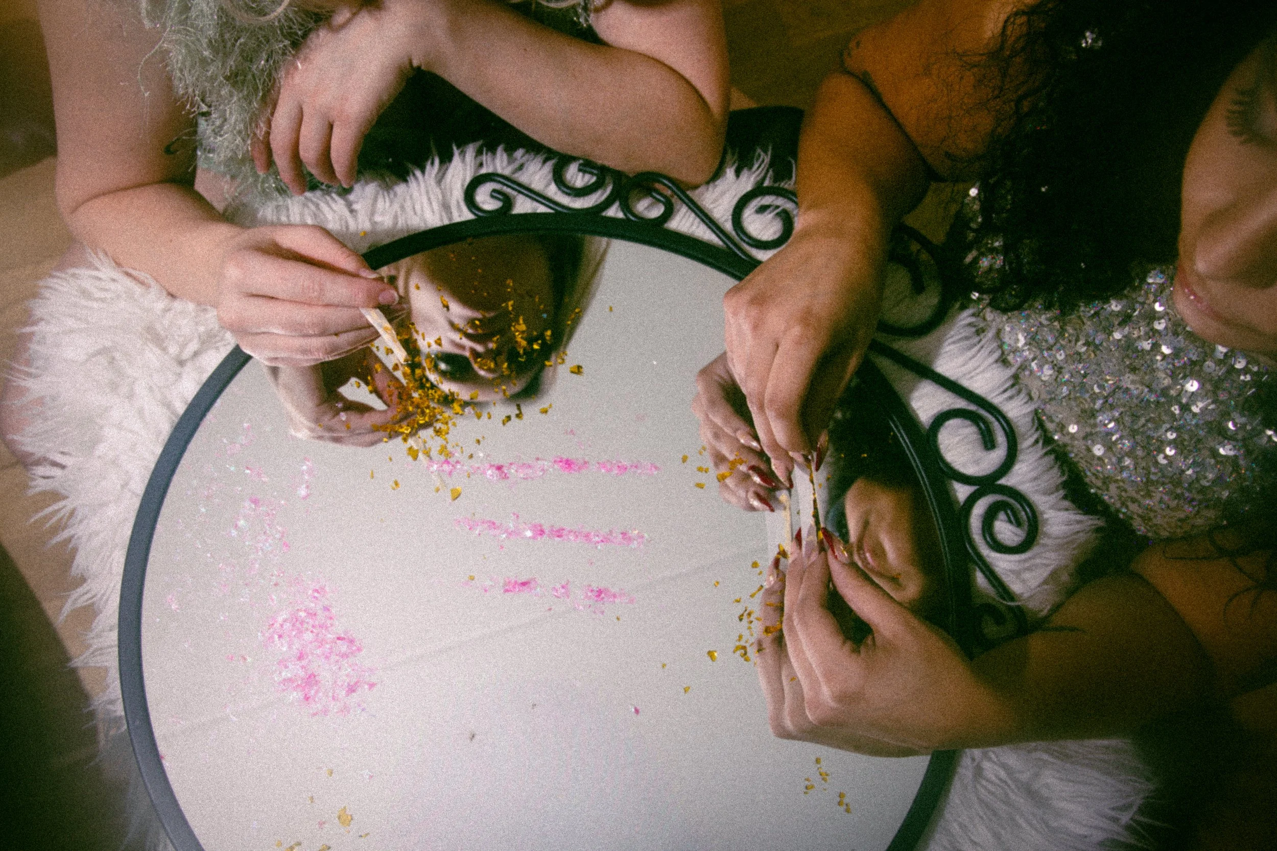Two women using makeup brushes to apply makeup in front of a round mirror with a reflective surface, placing gold glitter on their faces, with a white fluffy textured surface around the mirror.