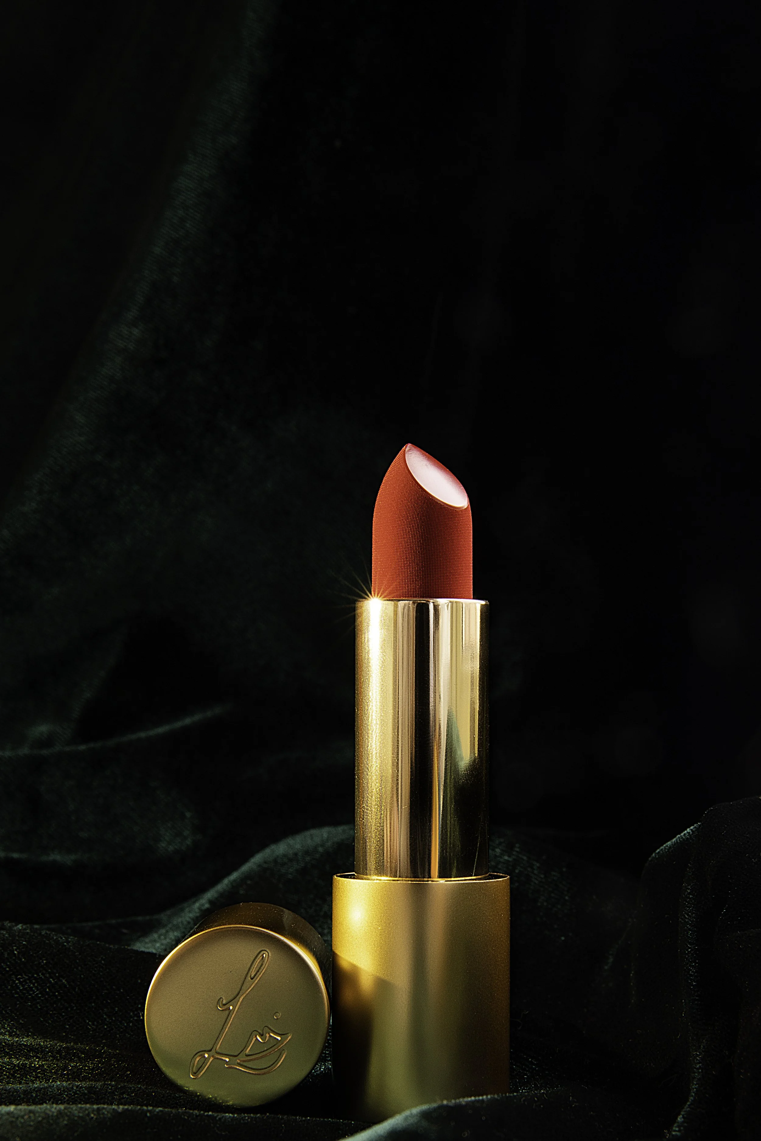 A red lipstick in a gold tube with a gold charm that has the signature 'L' on it. The lipstick is standing upright against a dark velvety background.
