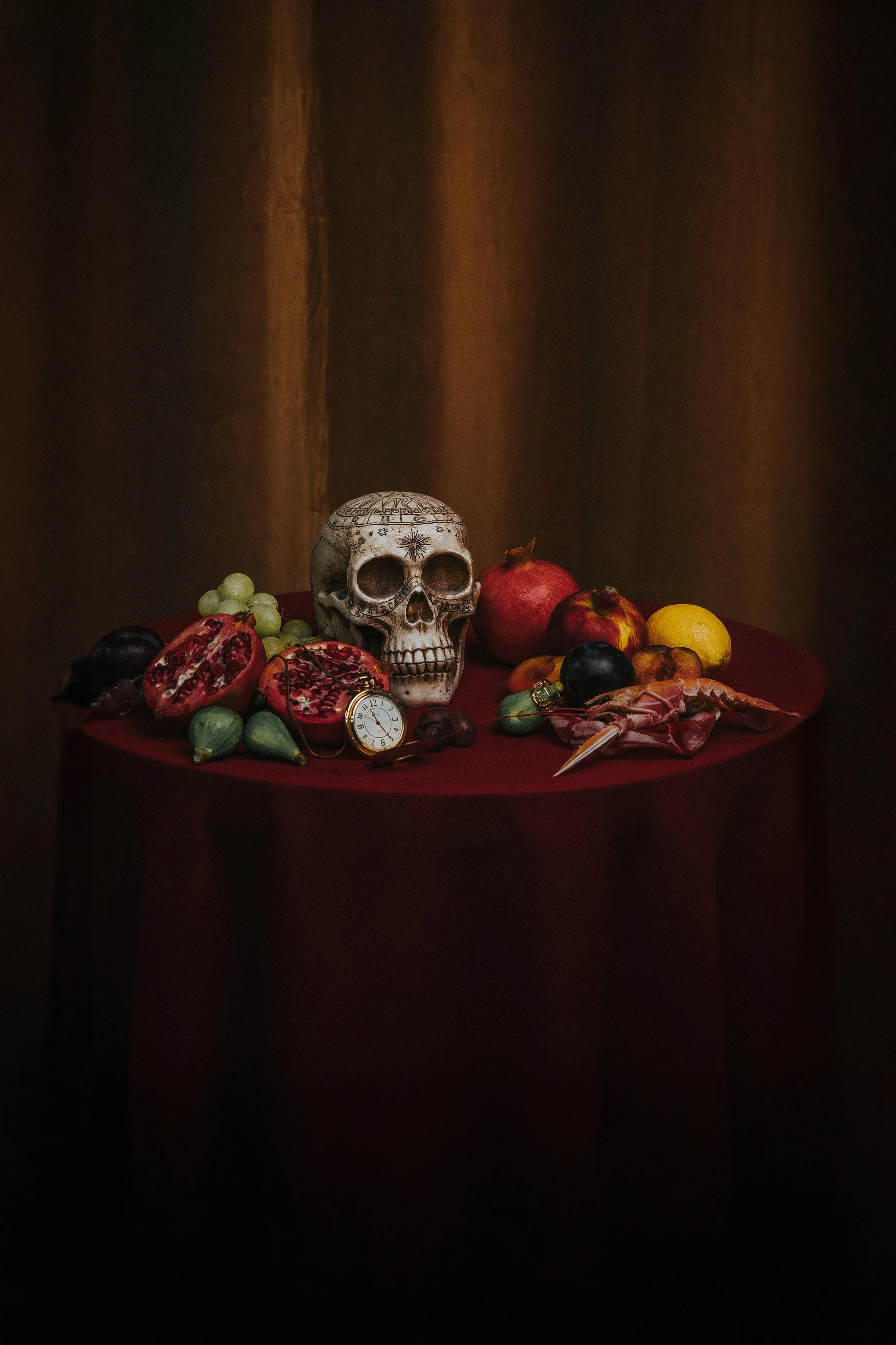 A still life arrangement featuring a white skull, pomegranates, grapes, plums, figs, and a pocket watch on a dark red tablecloth background.