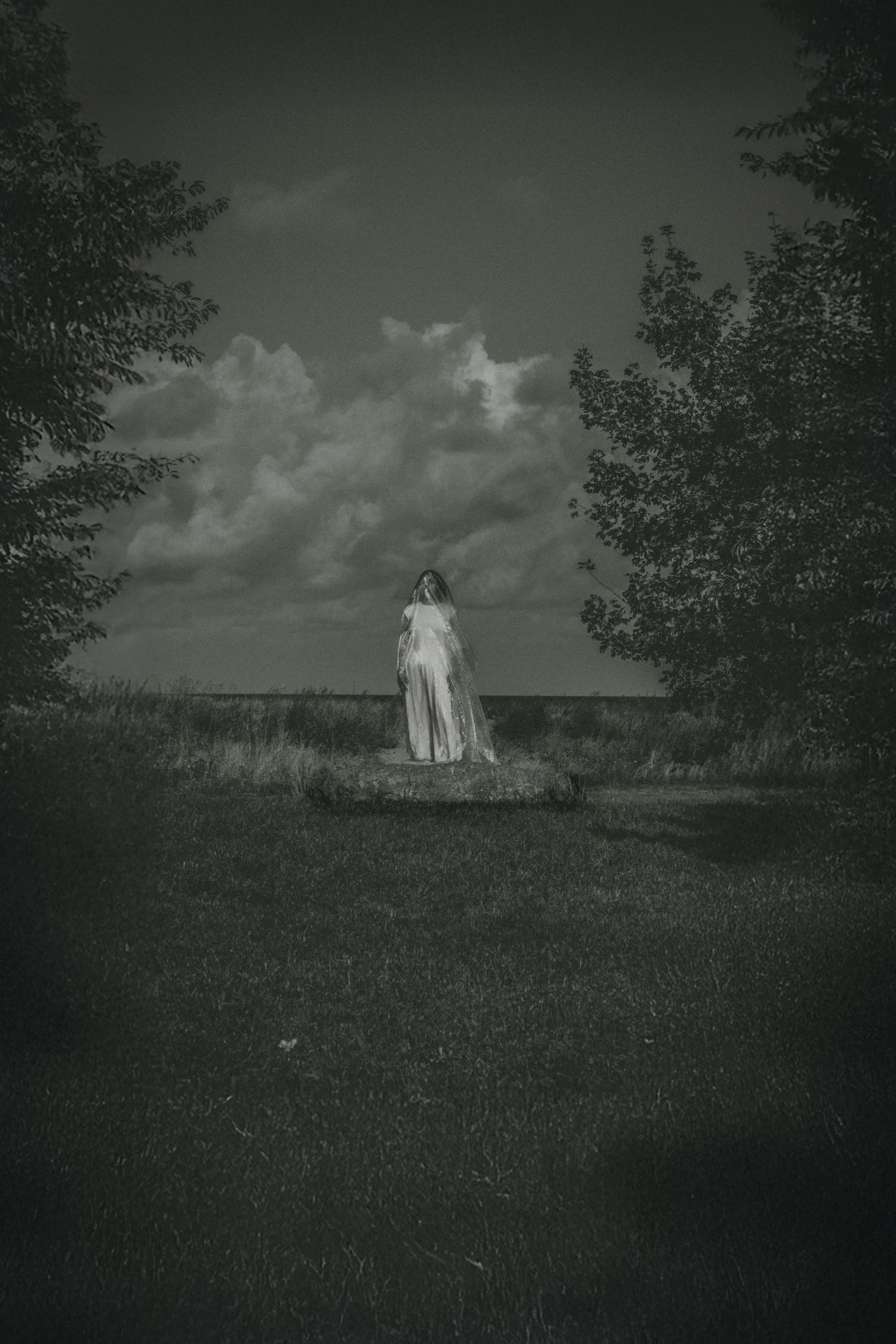 A black and white photo of a fountain with a sculpture of a cloaked figure at a park, framed by trees under a cloudy sky.