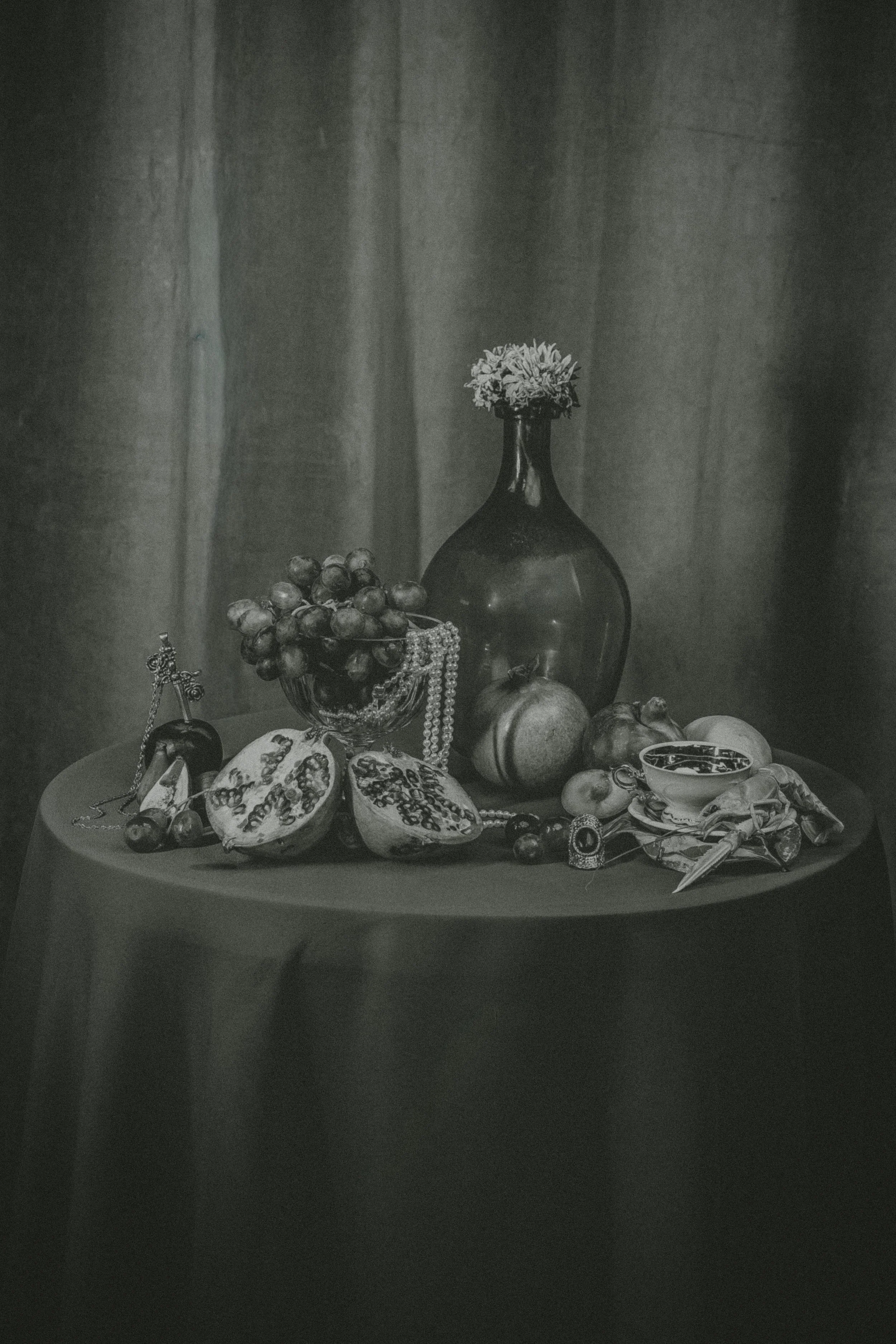 A still life composition of assorted fruits, jewelry, and decorative objects on a round table against a curtain backdrop.