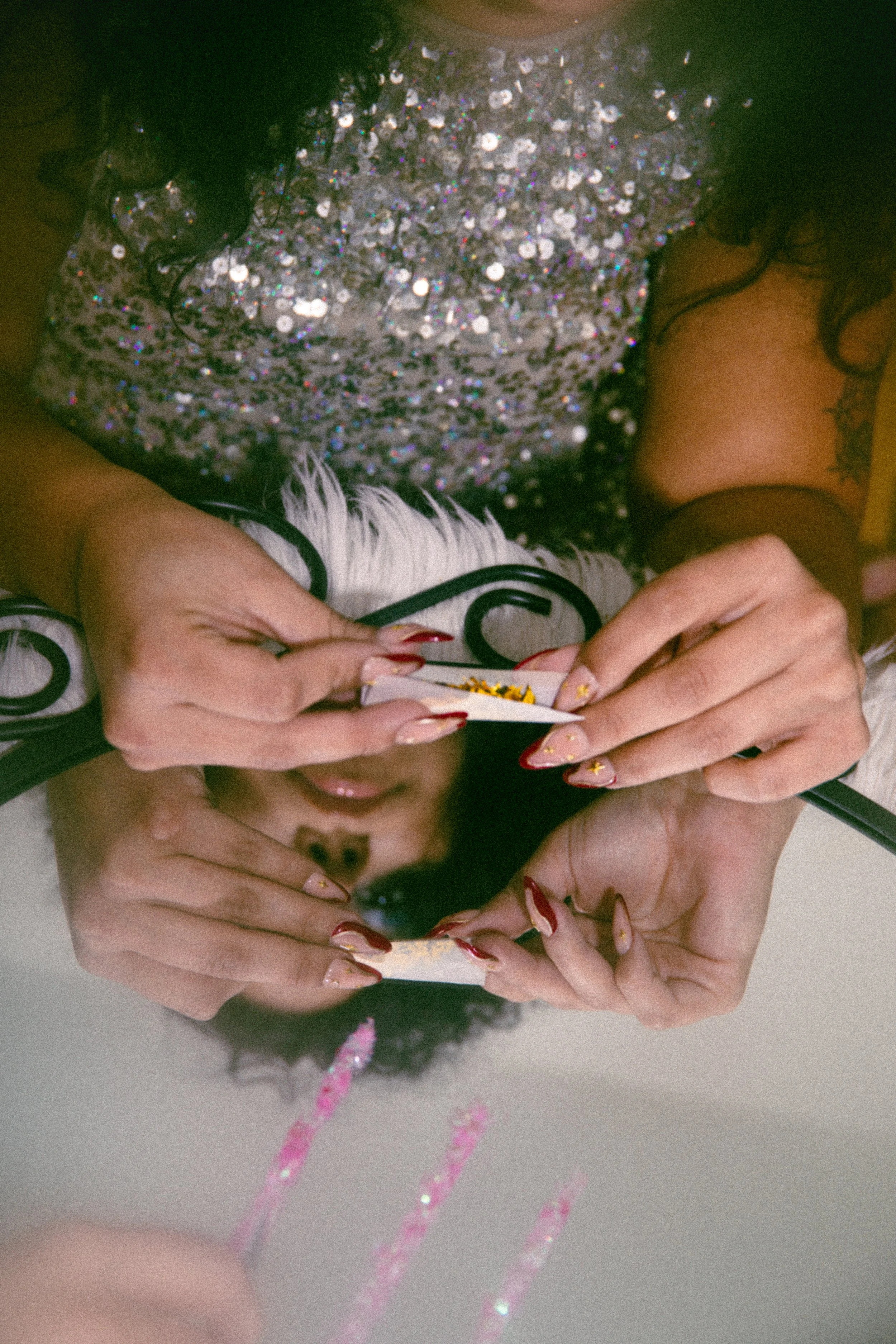Close-up of a person wearing a glittery silver dress with pink and black striped stockings, holding a pregnancy test with both hands. Their nails are painted with decorative designs. The person has a tattoo on their chest and long, dark curly hair.