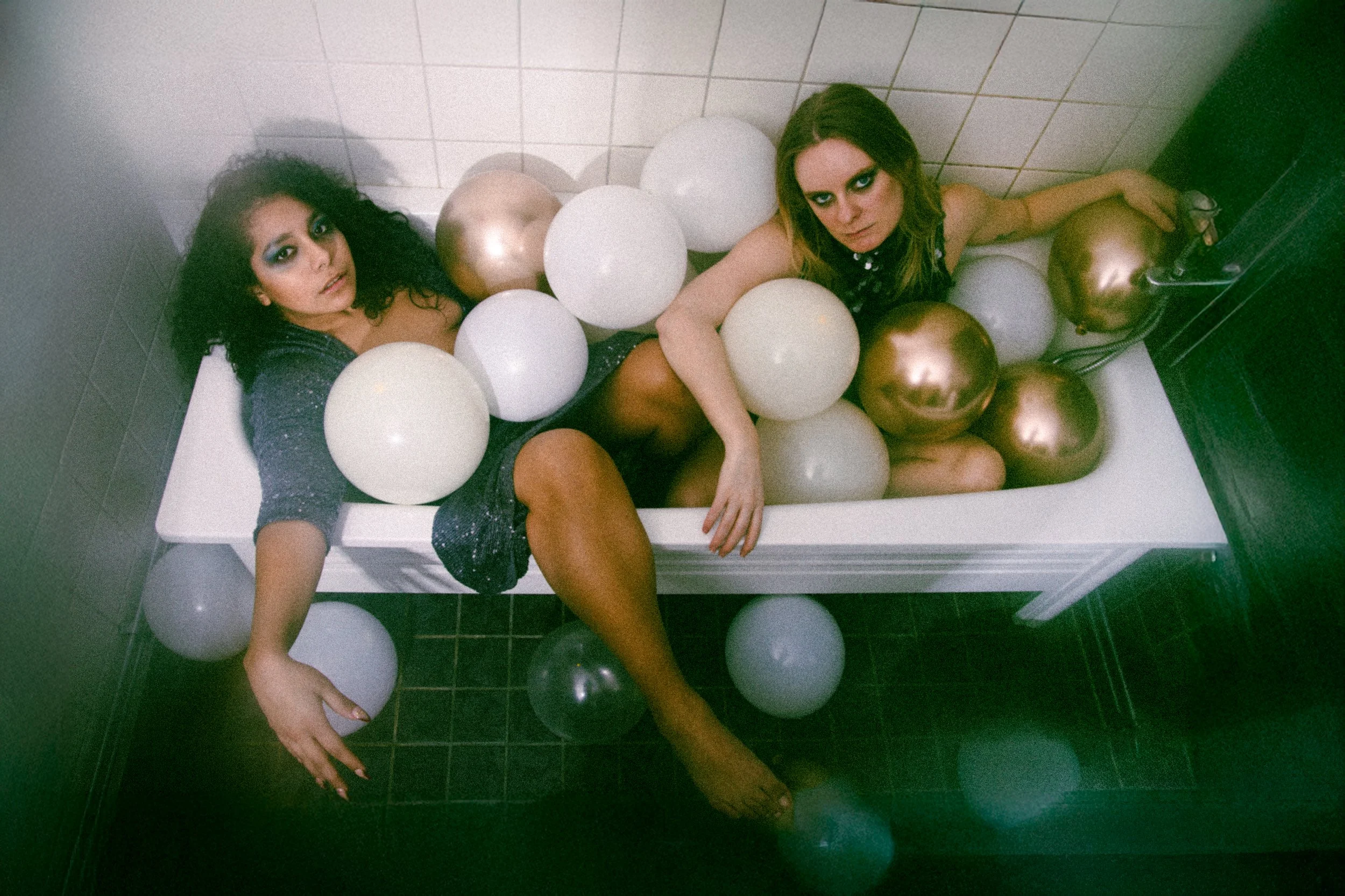 Two women are lying in a bathtub filled with white, gold, and silver balloons, with a white tiled bathroom wall in the background.