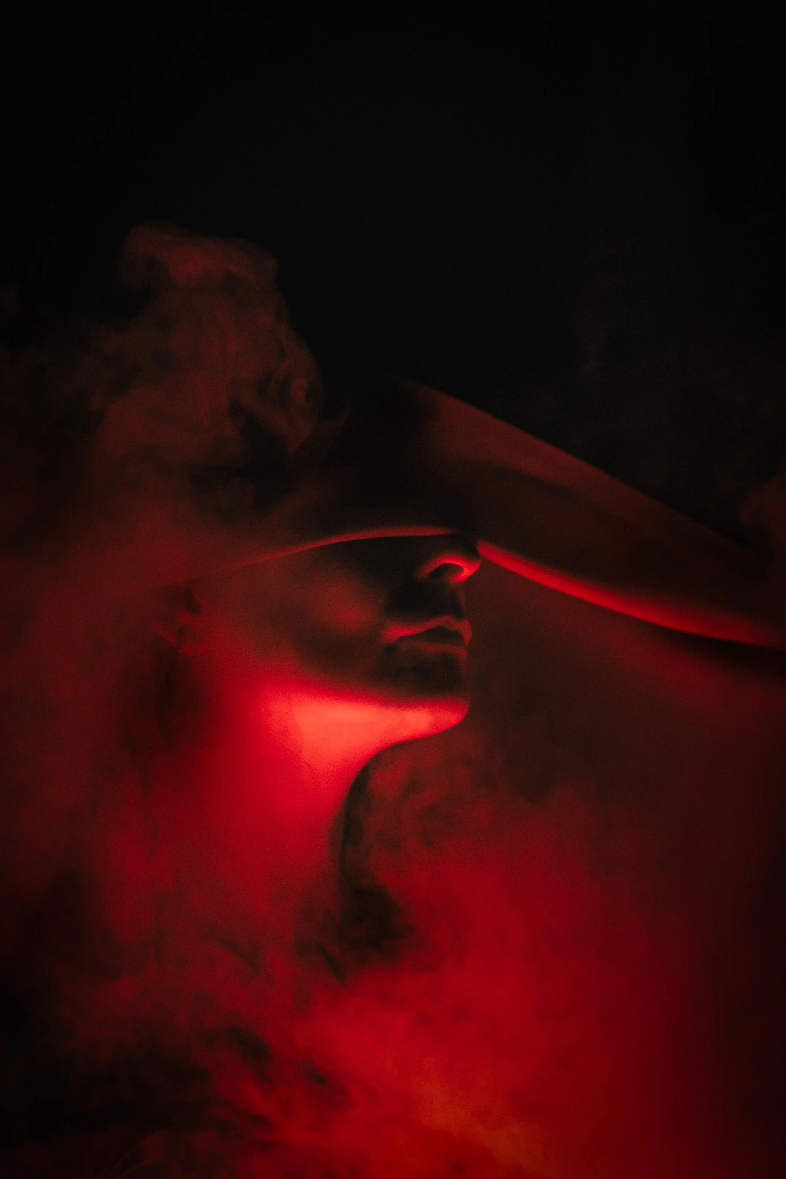 A photograph of a woman illuminated by red and orange lighting, with her eyes closed and her face draped in fabric, surrounded by smoke. Spooky, halloween vibe.